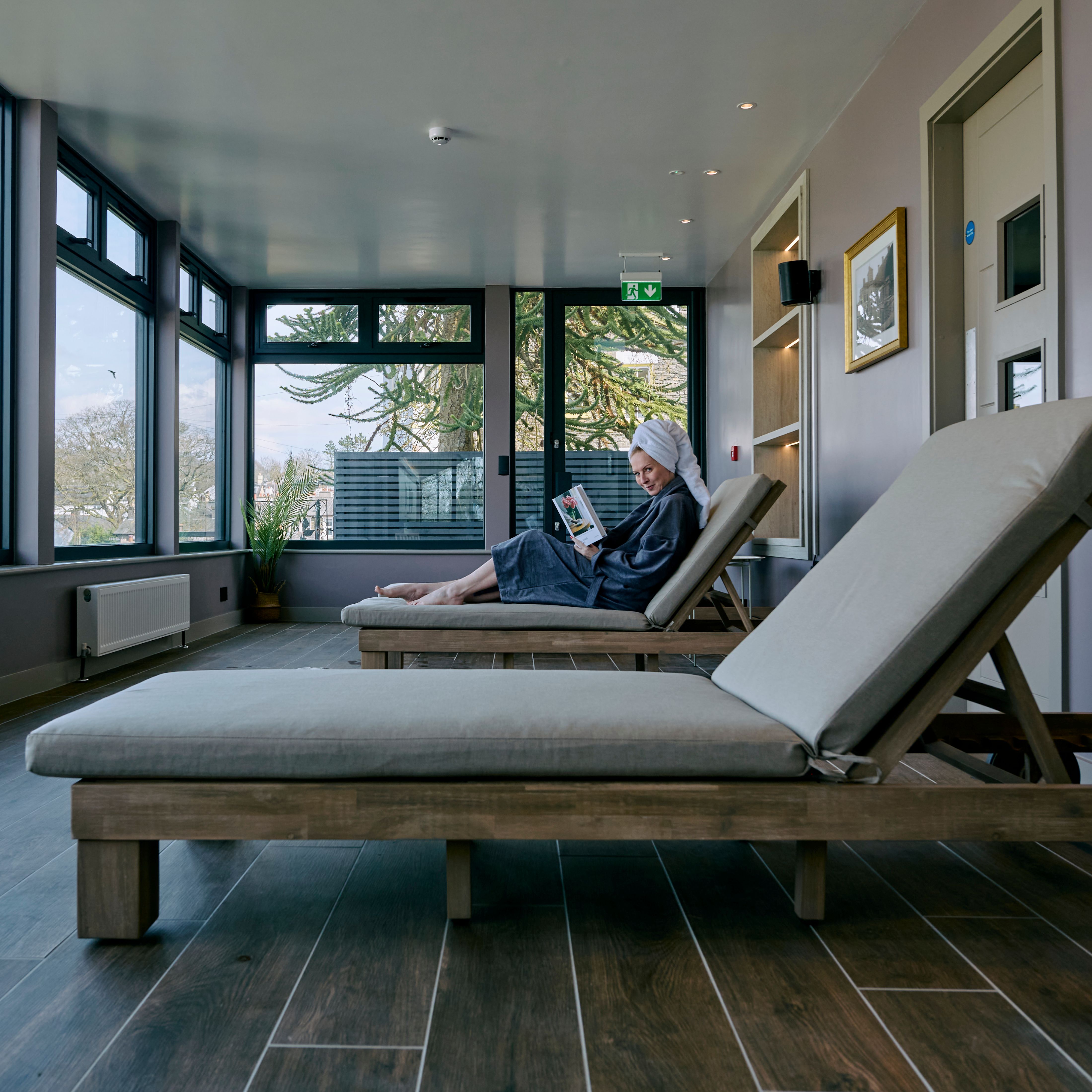 A woman in a robe and towel turban reads a book while lounging in a spa relaxation room with large windows overlooking a scenic view of rooftops and trees.