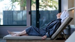 A woman in a robe and towel turban reclines on a cushioned lounge chair in a spa relaxation room, eyes closed and head tilted back in peaceful rest.