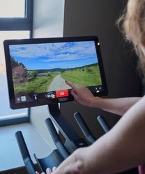 A woman interacts with a touchscreen on a cardio machine, selecting a virtual outdoor trail with a scenic path, grassy fields, and blue sky.