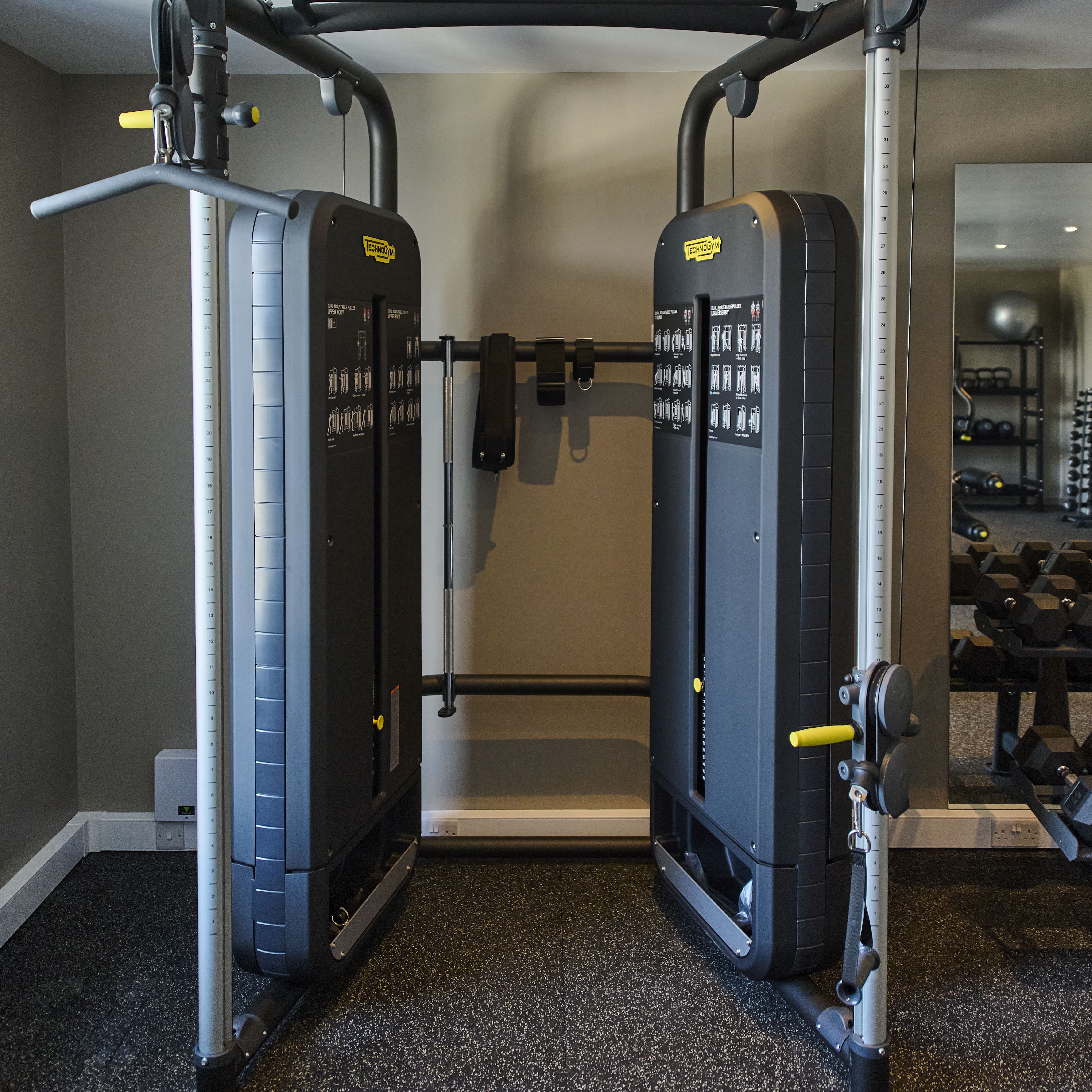 A dual adjustable pulley machine in a gym, with illustrated exercise guides on each weight stack and a pull-up bar overhead, set against a neutral wall.