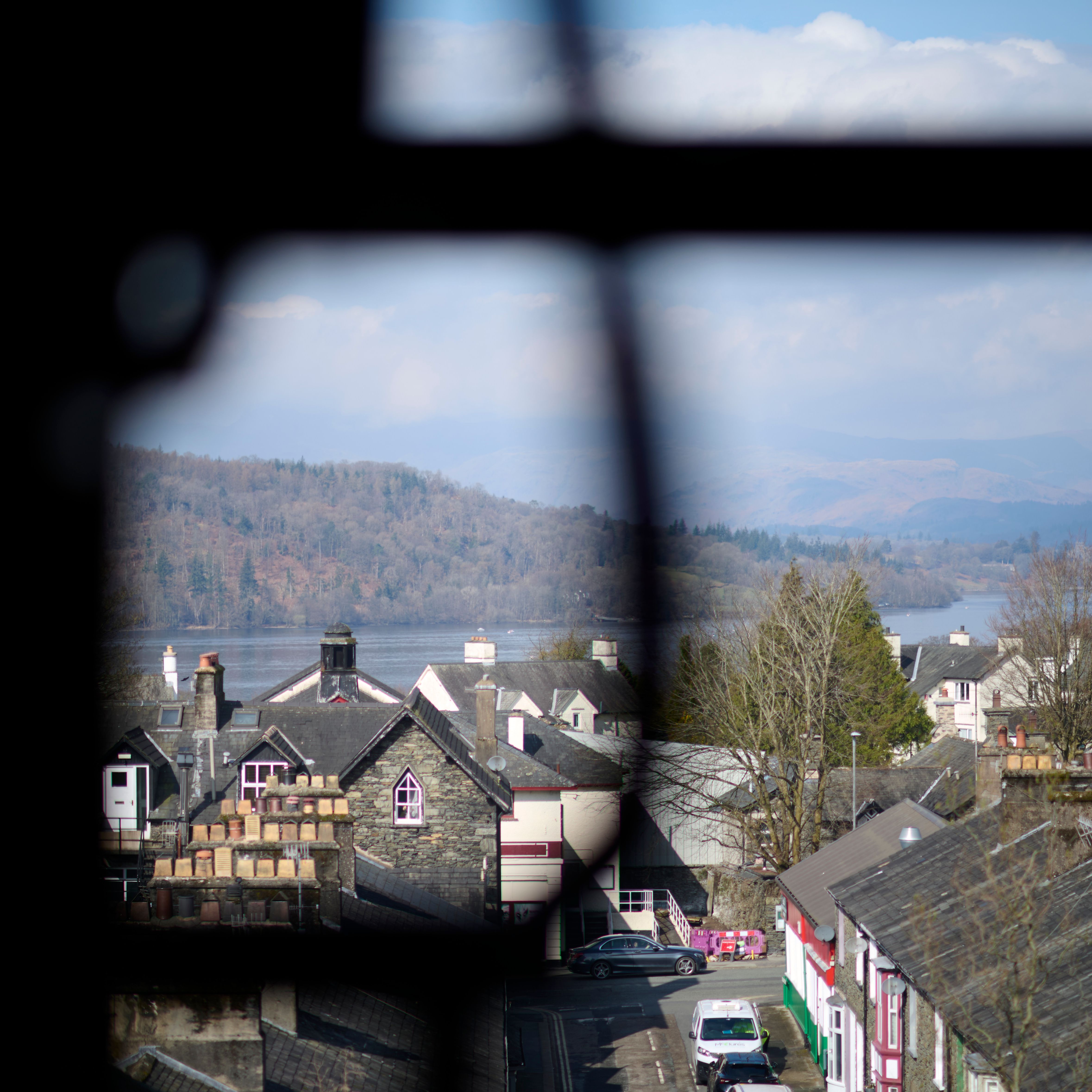 Scenic view through a window overlooking rooftops of a quaint town, with a lake and tree-covered hills in the background under a partly cloudy sky.