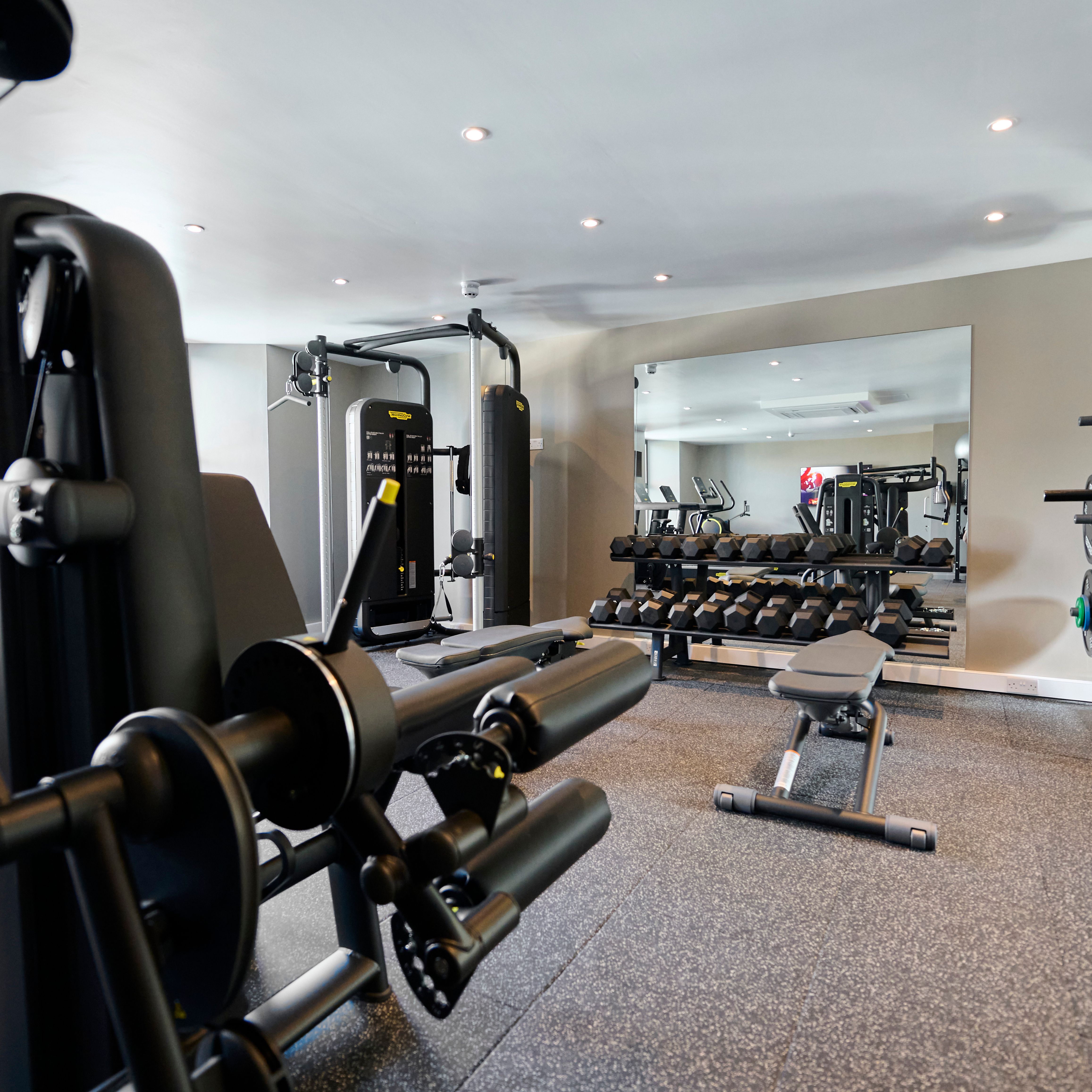 A modern gym with various strength training machines, free weights, benches, and a Smith machine, all reflected in a large wall mirror.
