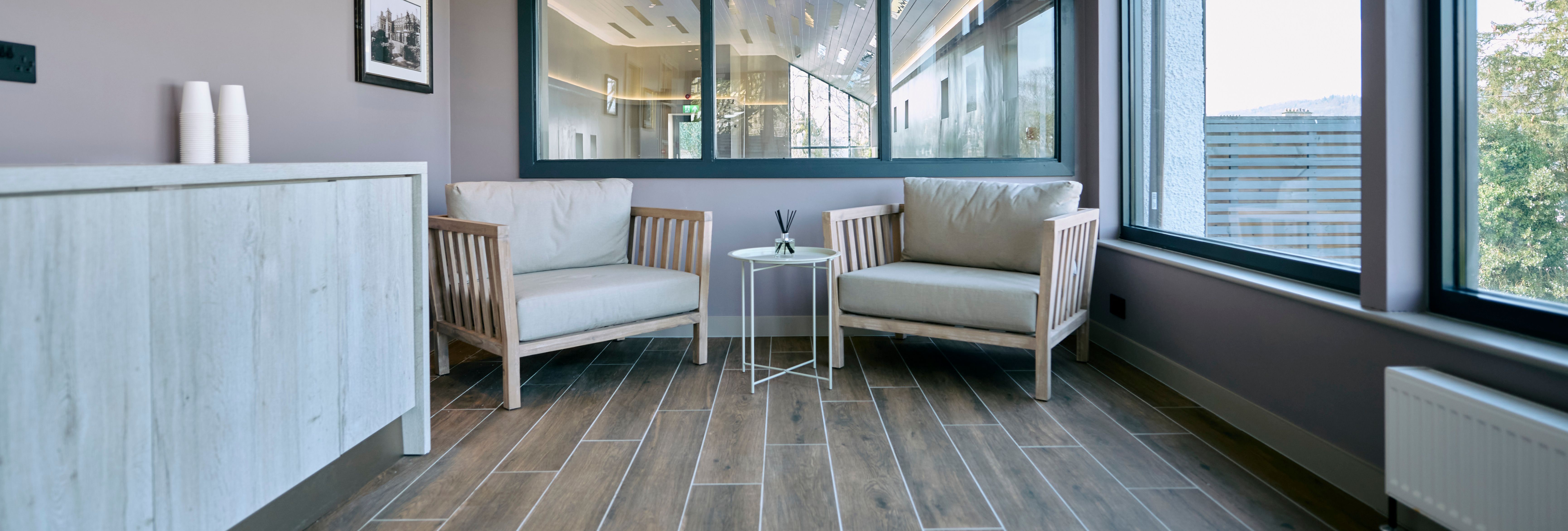 A cozy spa lounge area with two cushioned wooden chairs, a small table with a reed diffuser, large windows, and wood-style flooring.