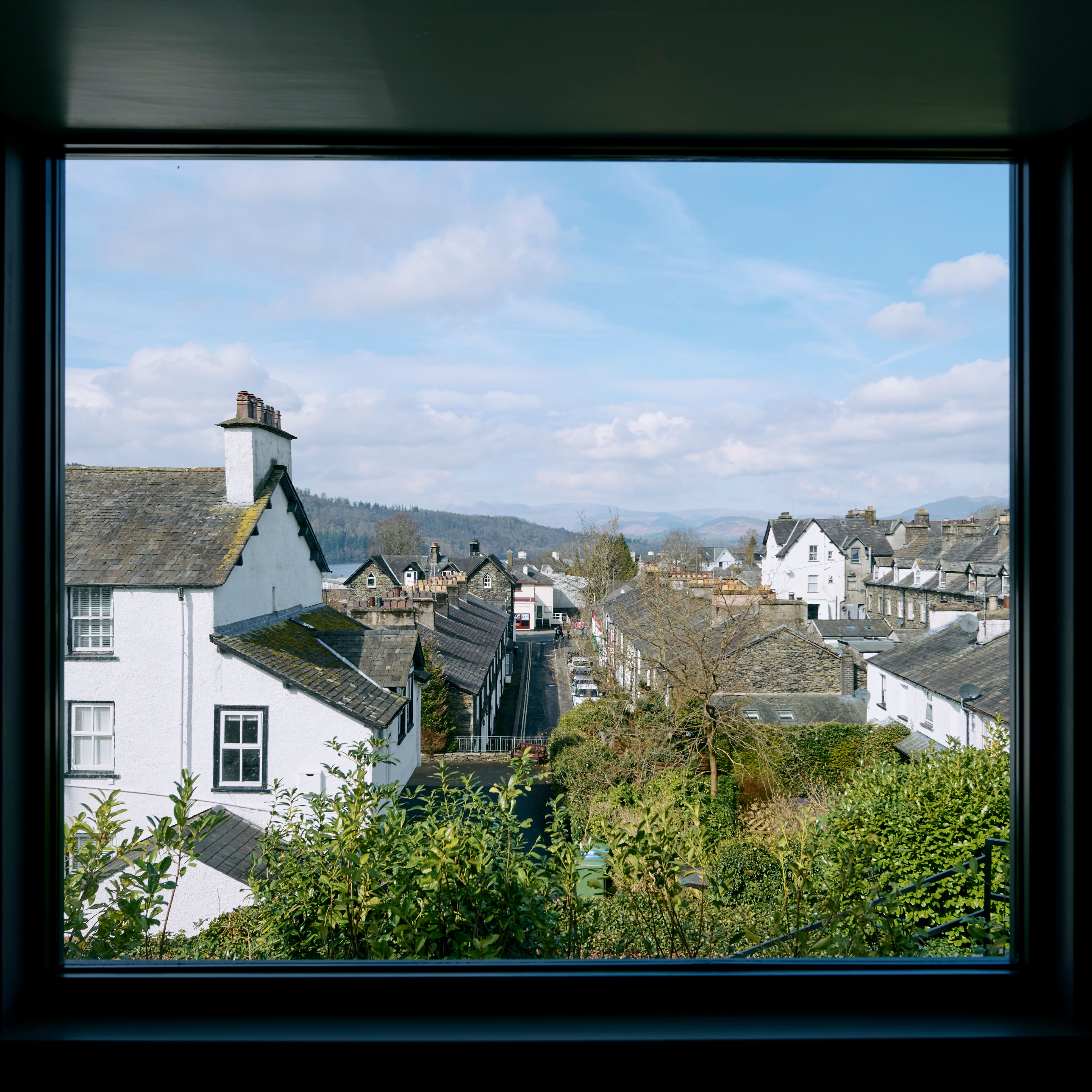 View through large windows overlooking a charming town with white and stone buildings, greenery, and distant hills under a bright blue sky with clouds.