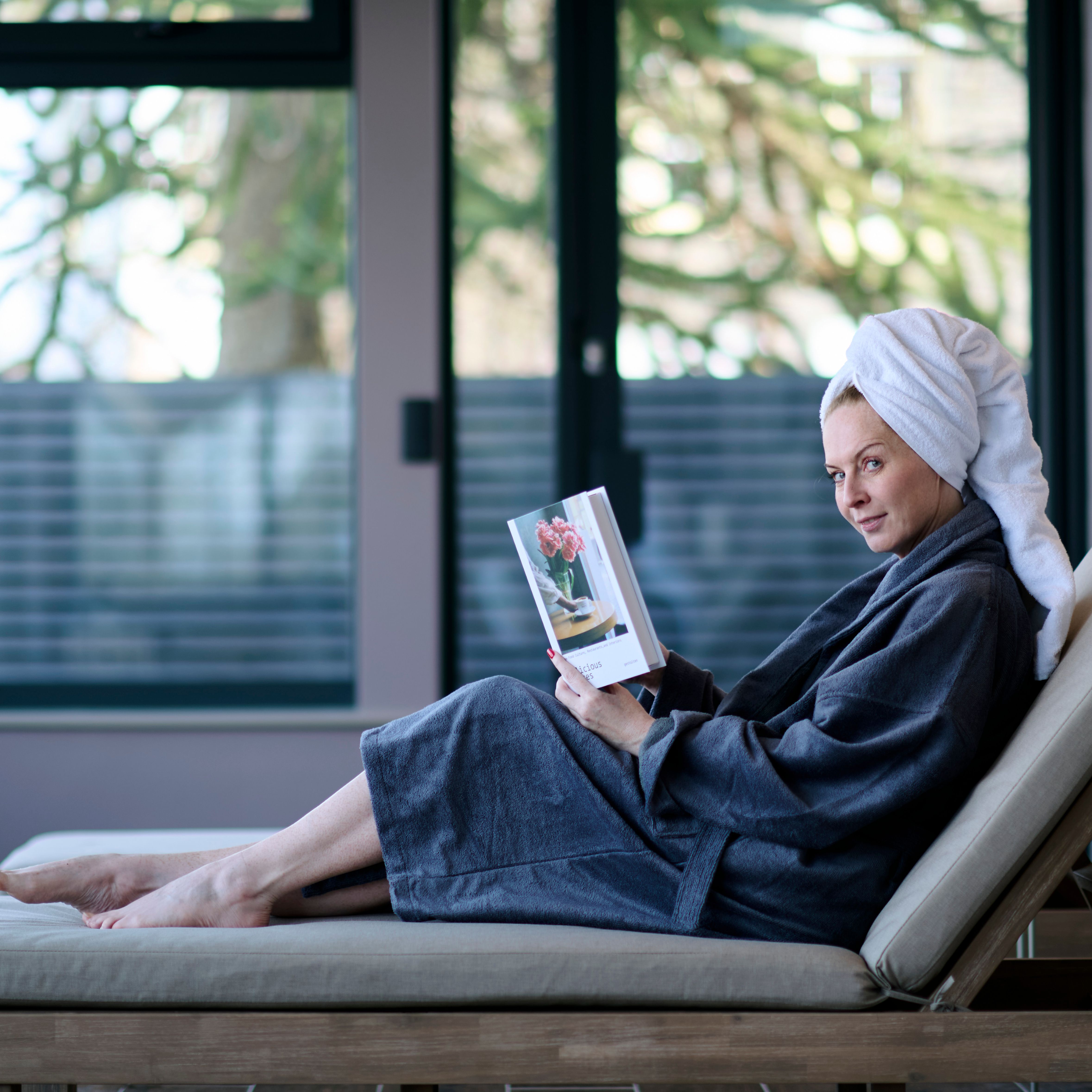 A woman in a bathrobe and towel turban lounges on a spa chair, smiling slightly while reading a book titled 
