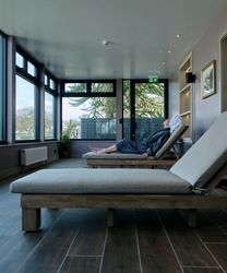 A woman in a robe and towel turban relaxes on a lounge chair in a spa relaxation room with large windows overlooking rooftops and trees.