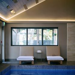 Two cushioned lounge chairs with white towels face an indoor pool, reflected in the calm water, beneath a sloped ceiling with soft ambient lighting.