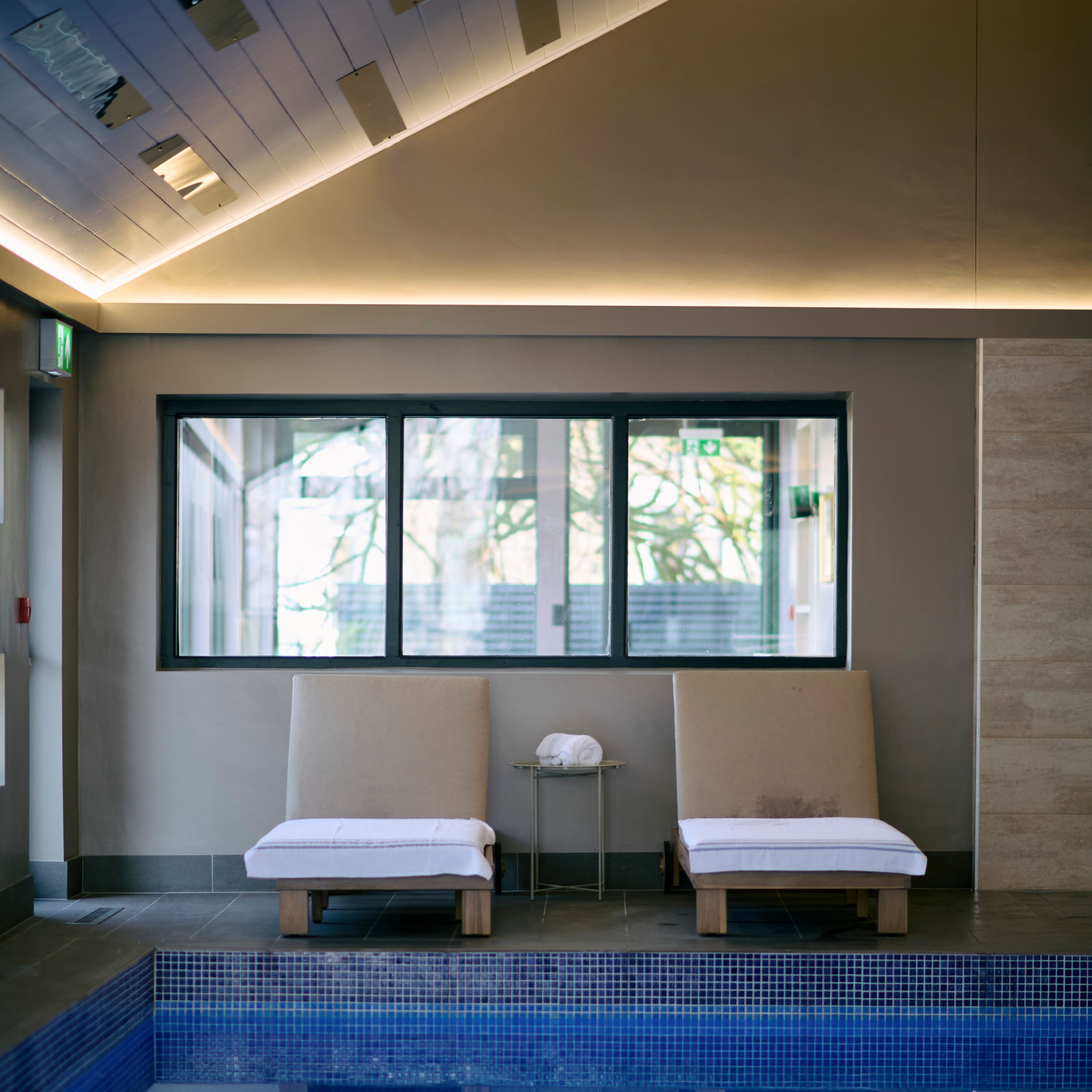 Two cushioned lounge chairs with white towels face an indoor pool, reflected in the calm water, beneath a sloped ceiling with soft ambient lighting.