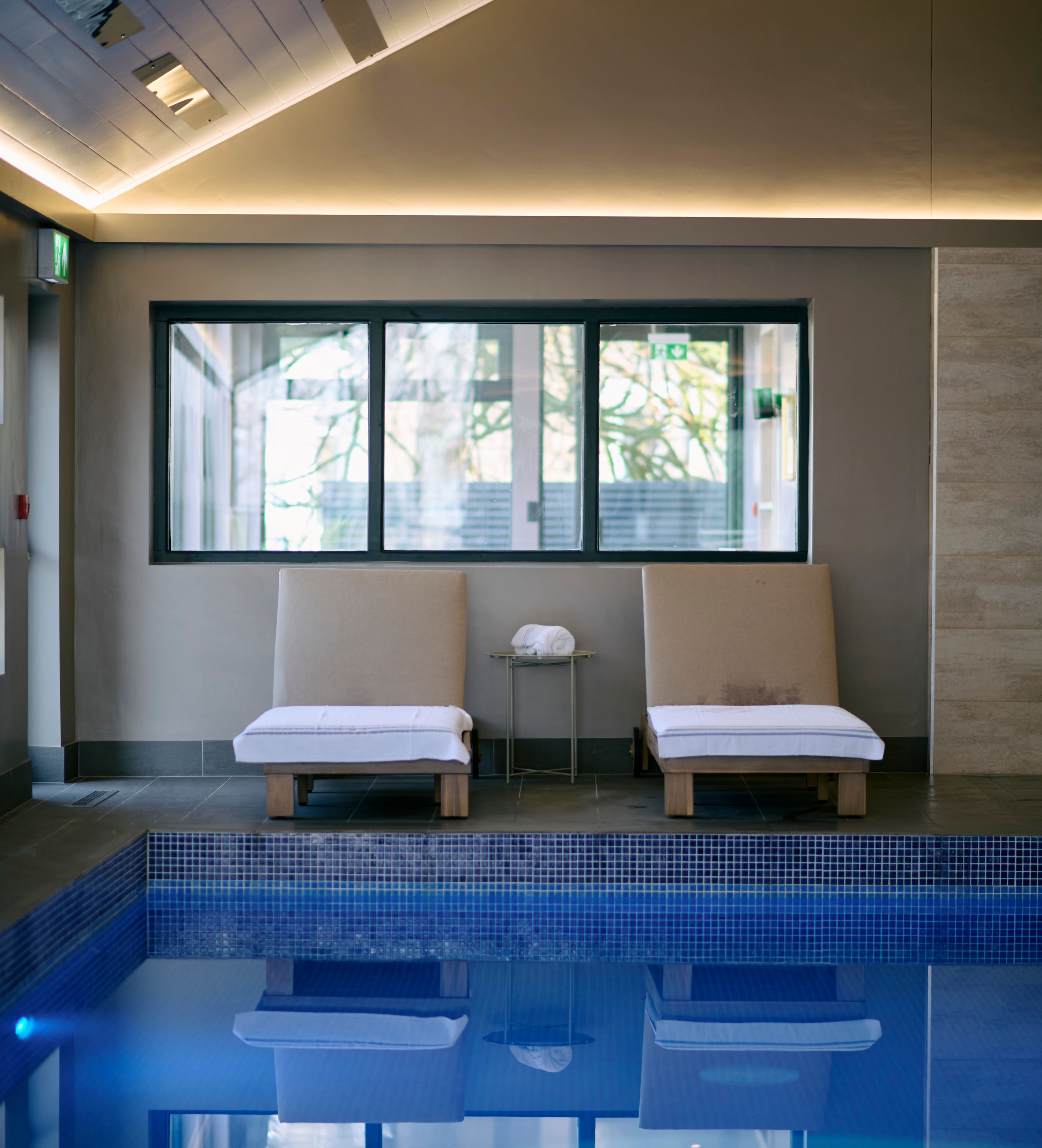 Two cushioned lounge chairs with white towels face an indoor pool, reflected in the calm water, beneath a sloped ceiling with soft ambient lighting.