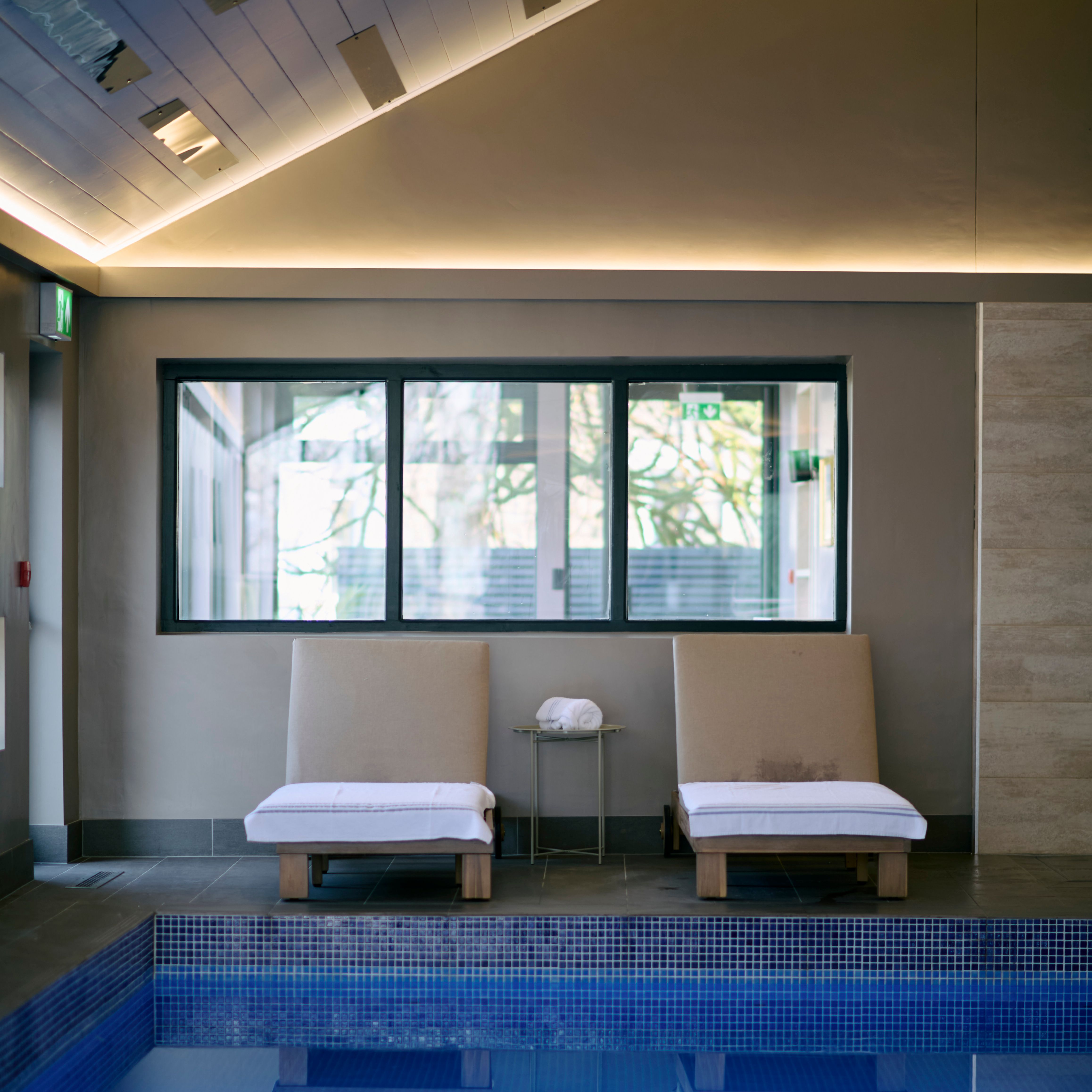 Two cushioned lounge chairs with white towels face an indoor pool, reflected in the calm water, beneath a sloped ceiling with soft ambient lighting.
