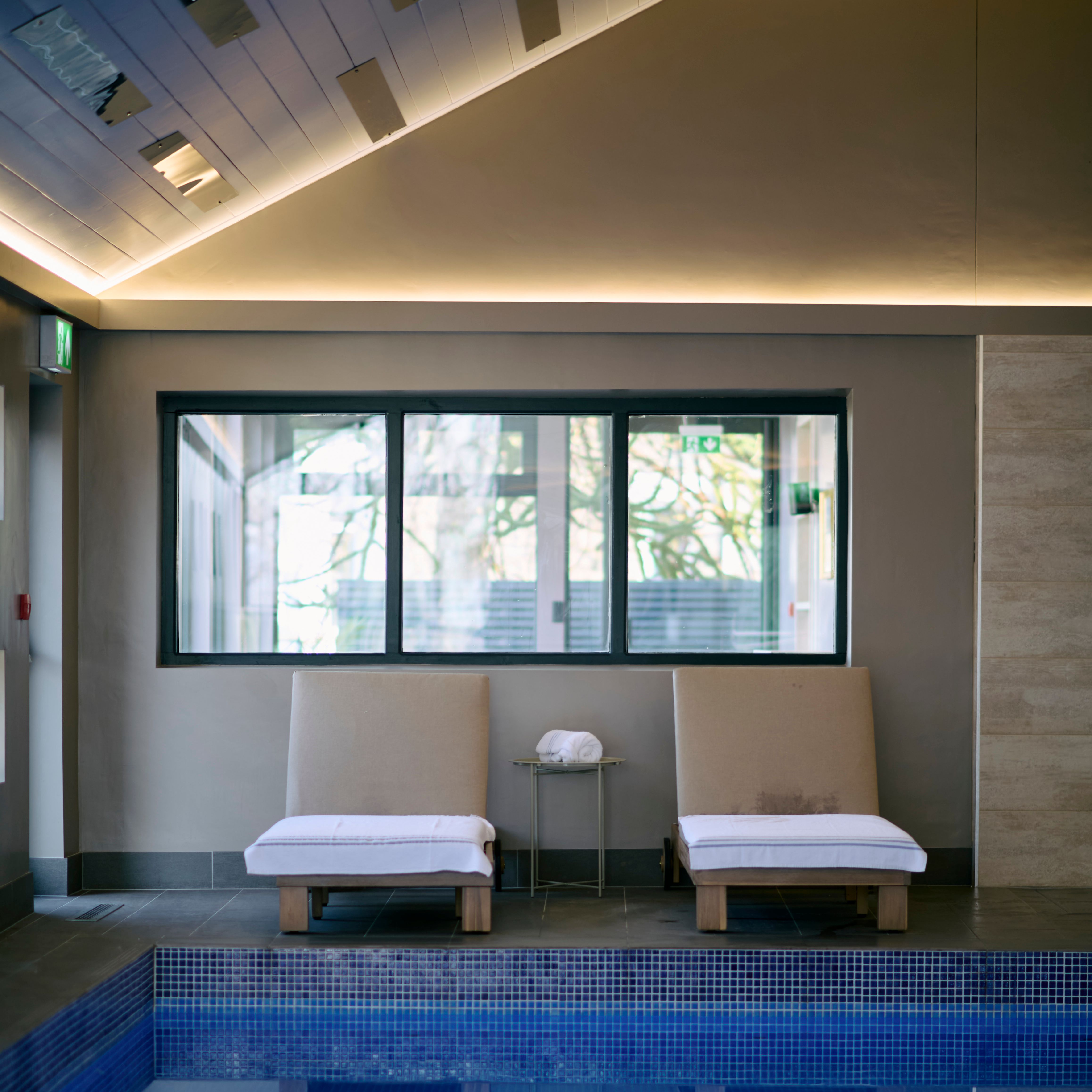 Two cushioned lounge chairs with white towels face an indoor pool, reflected in the calm water, beneath a sloped ceiling with soft ambient lighting.
