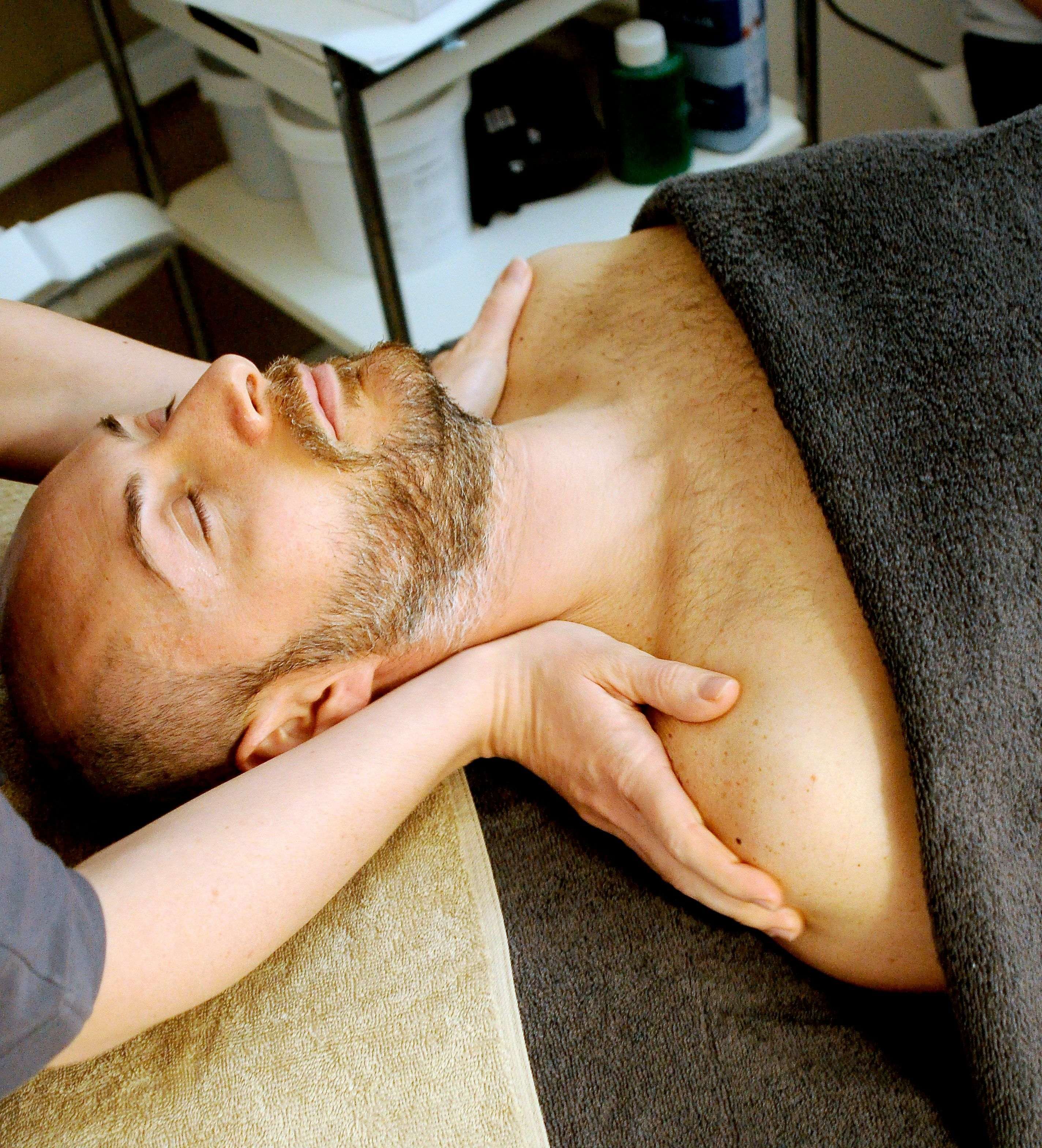 A man receiving a comforting massage lies on a massage bed, with the masseuse applying pressure to the collarbones.