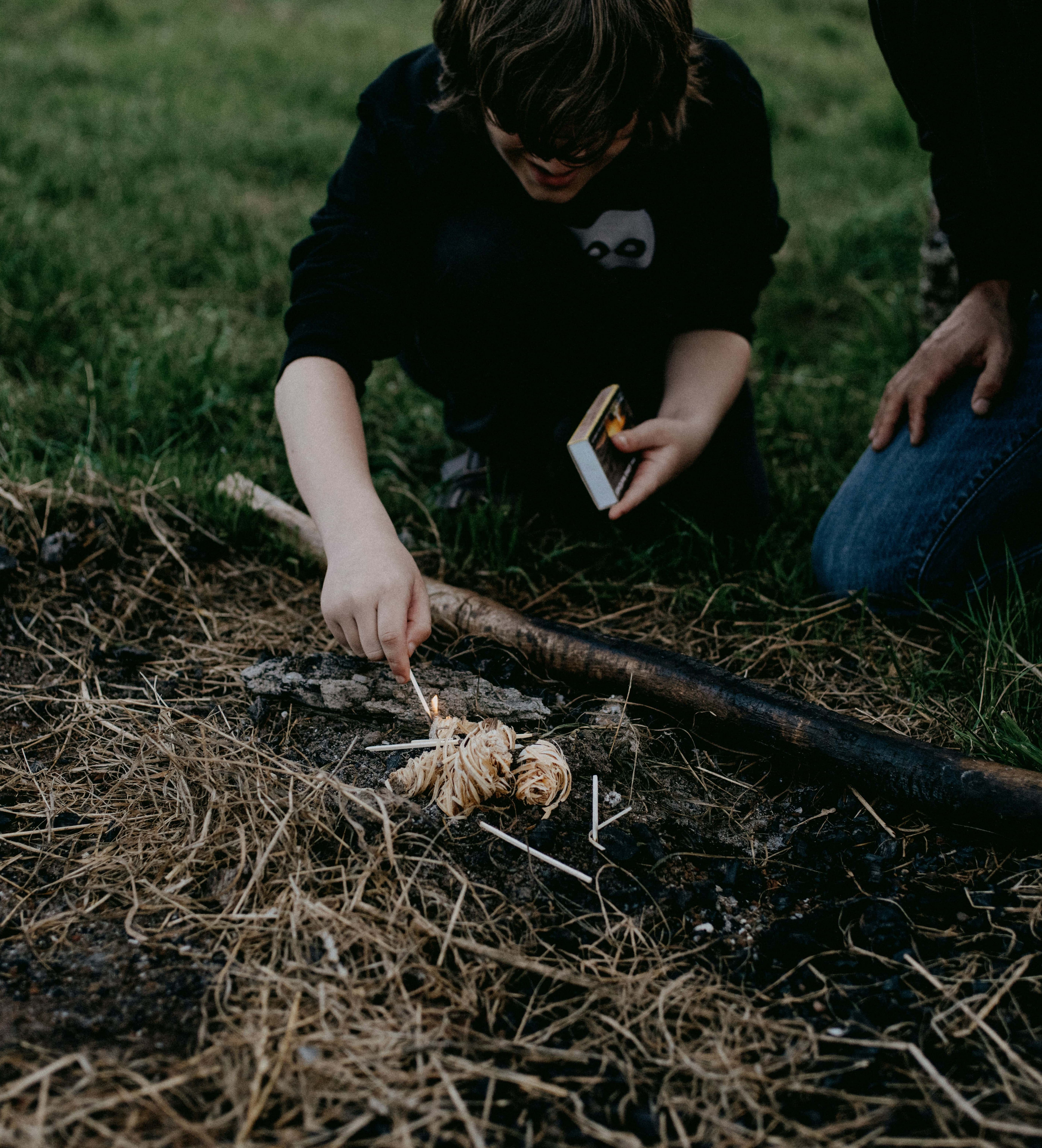A father and his son are engaging in bushcraft activities. The child is lighting some kindling on fire to practise his fire making skills.