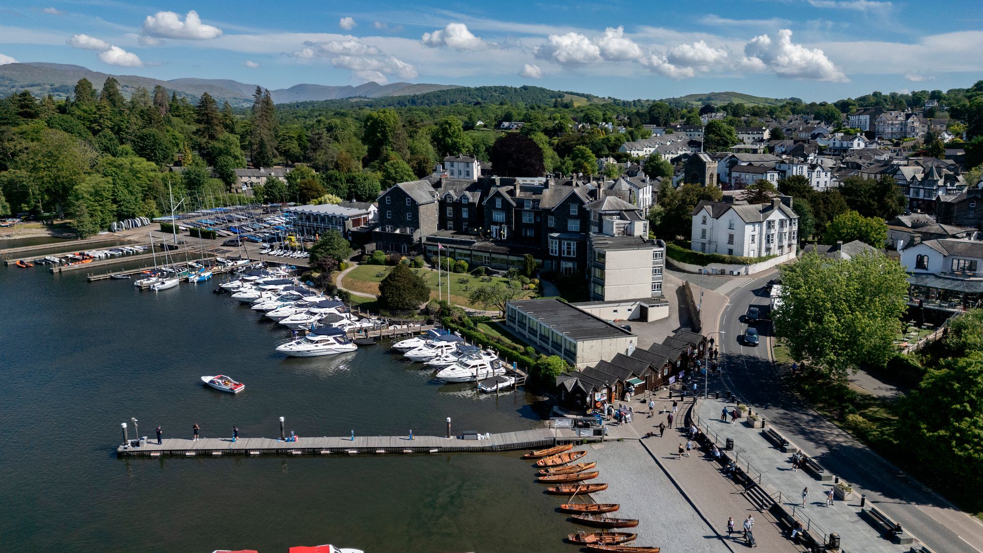 Aerial view of Bowness-on-Windermere marina with boats docked on the lake, surrounded by town buildings, trees, and distant hills under a blue sky.