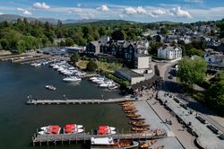 Aerial view of Bowness-on-Windermere marina with boats docked on the lake, surrounded by town buildings, trees, and distant hills under a blue sky.