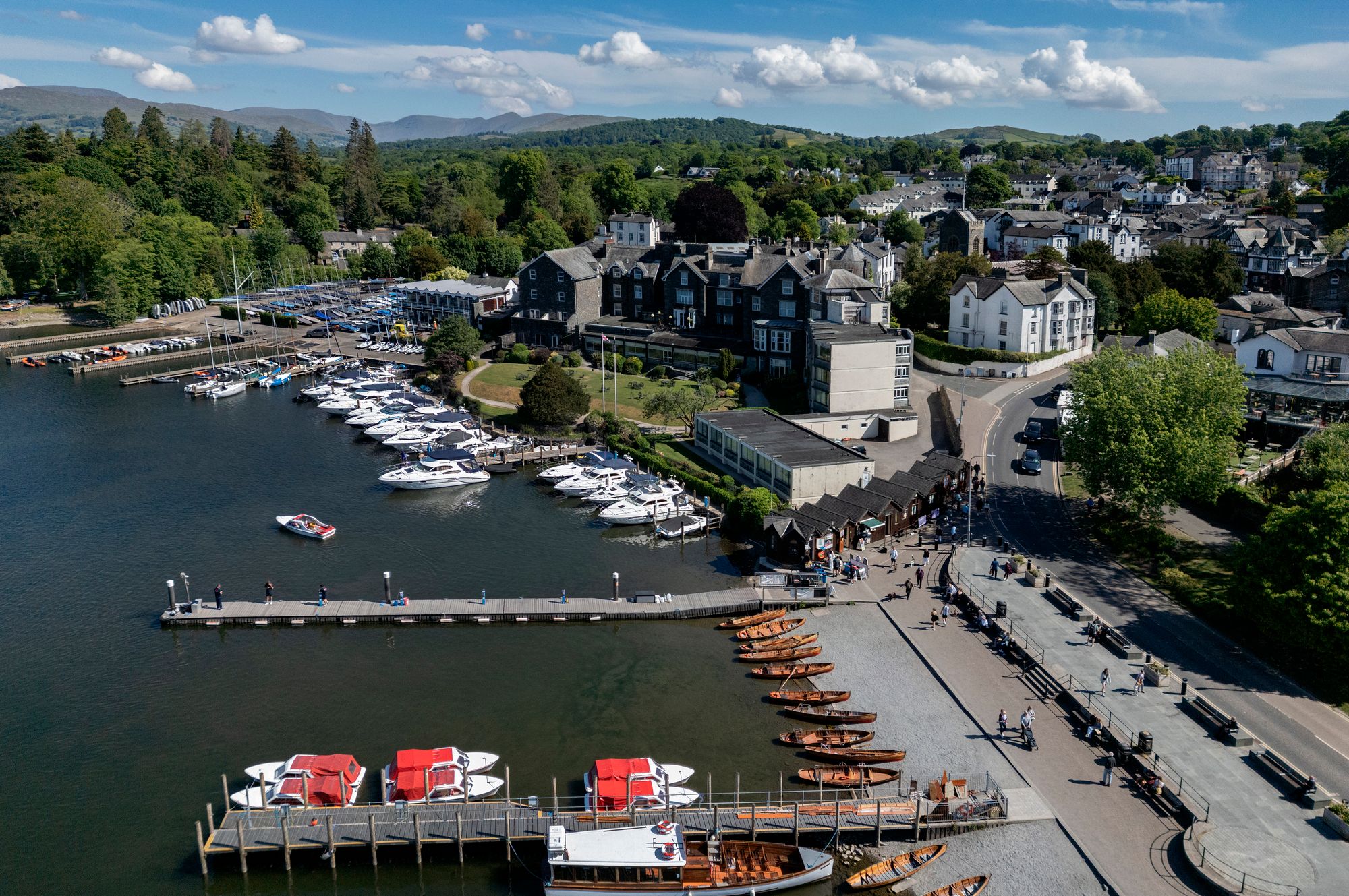 Aerial view of Bowness-on-Windermere marina with boats docked on the lake, surrounded by town buildings, trees, and distant hills under a blue sky.