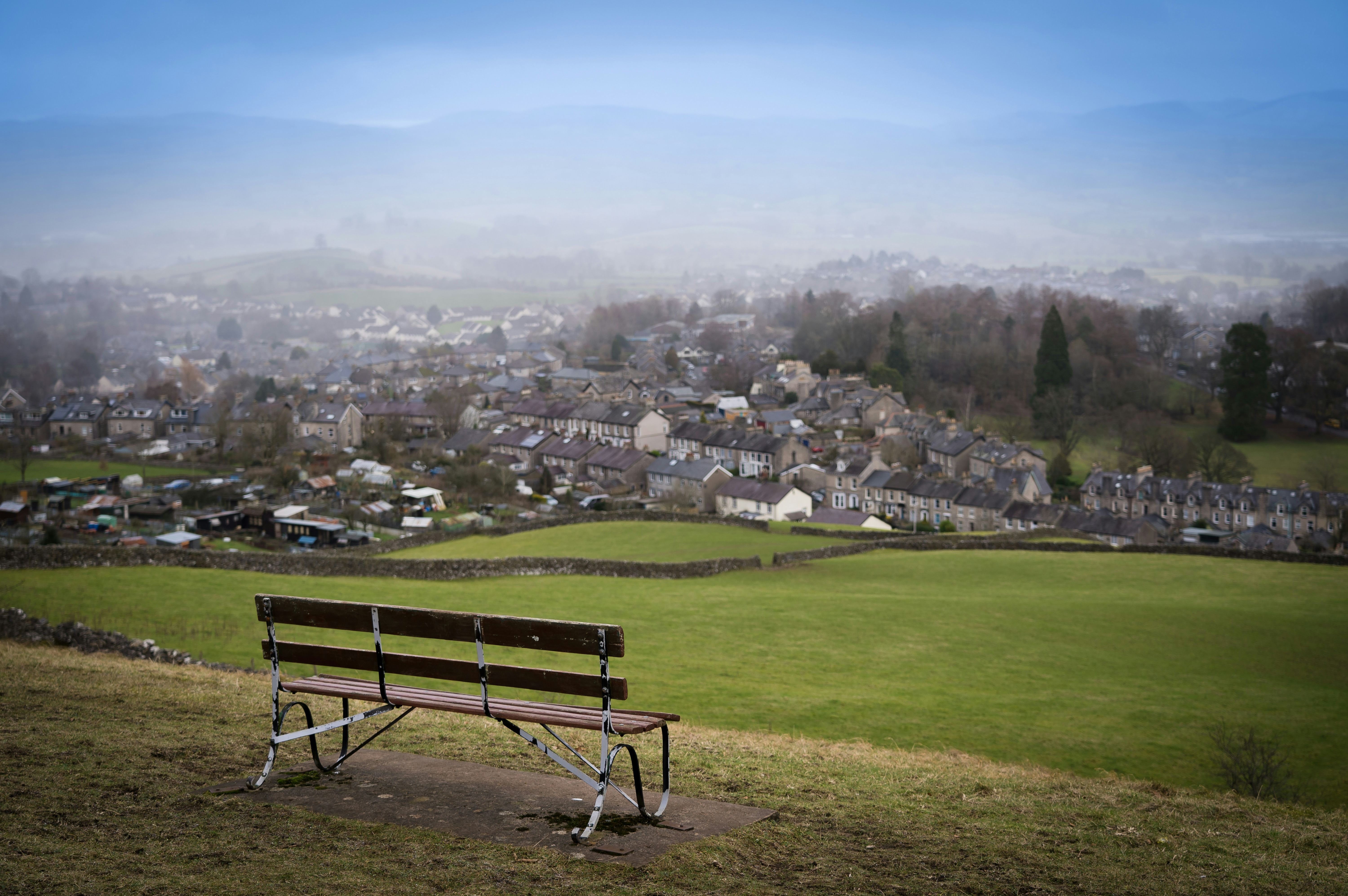 Empty bench on a grassy hill overlooking a small town with houses and fields under a misty sky