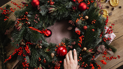 Hand decorating a festive holiday wreath with red ornaments and berries on a wooden table.