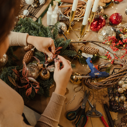 Person crafting a festive holiday wreath with ornaments, pinecones, and greenery on a decorated table