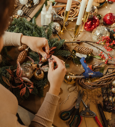 Person crafting a festive holiday wreath with ornaments, pinecones, and greenery on a decorated table