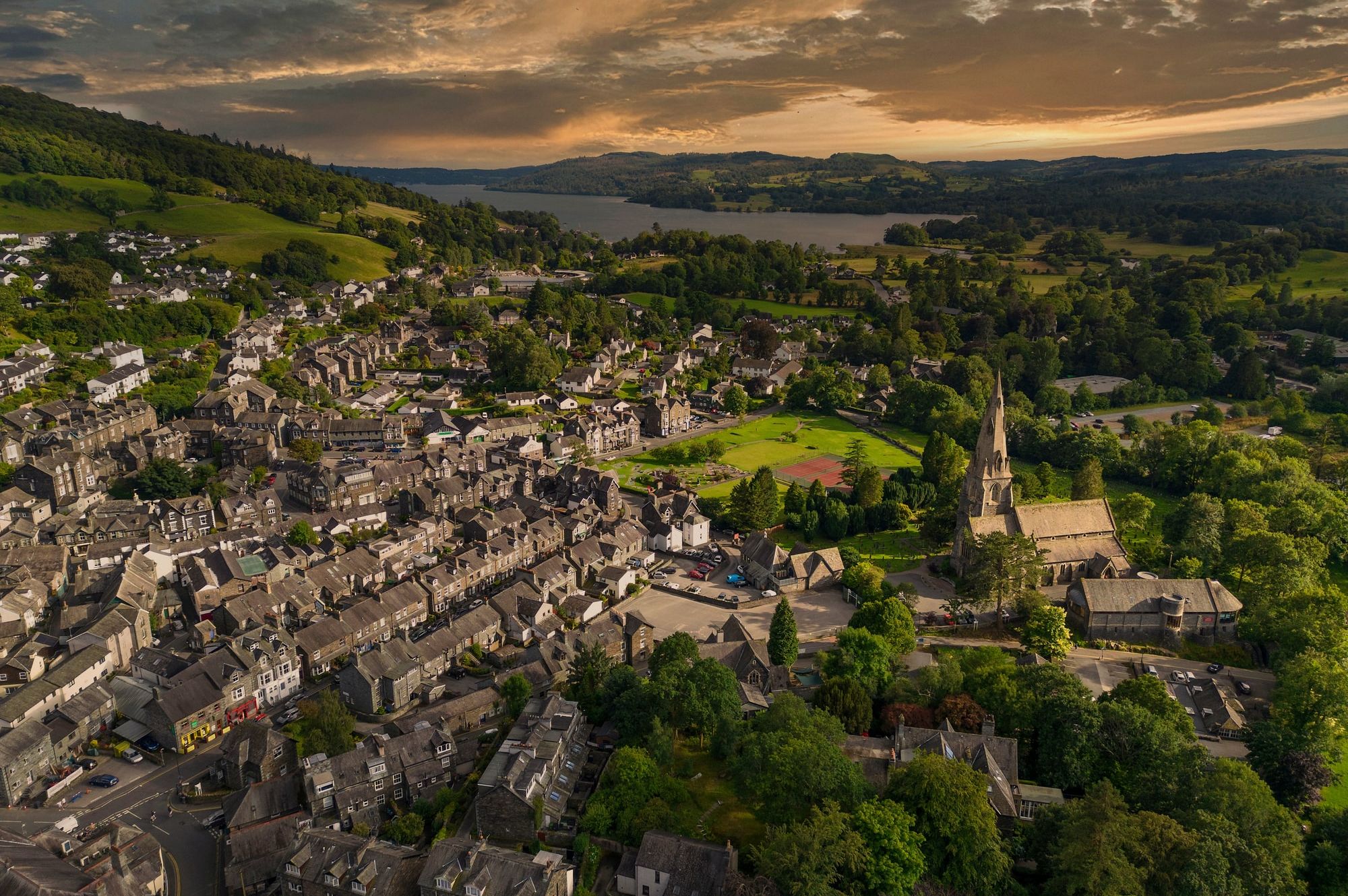 Aerial view of Ambleside in the Lake District, showing stone buildings, a church with a tall spire, green hills, and Lake Windermere at sunset.