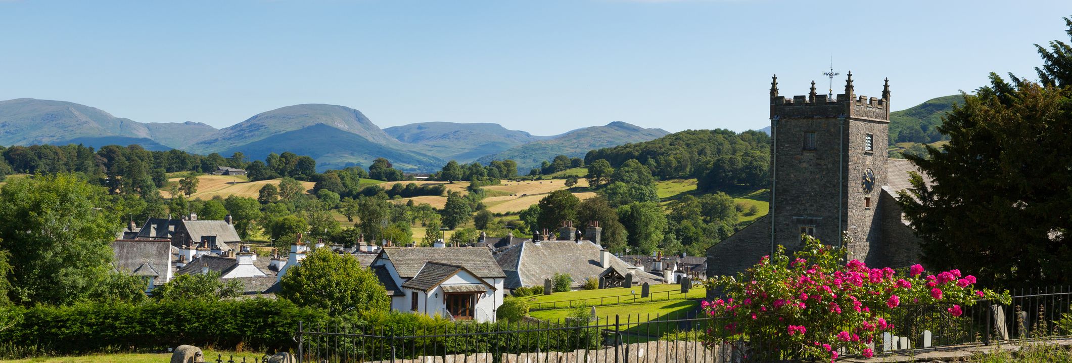 Hawkshead village in the Lake District, showing a stone church tower, rooftops, green fields, and distant hills under a bright blue sky.