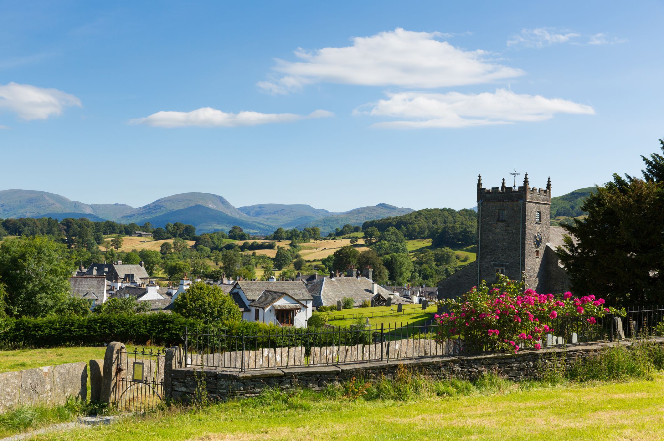 Hawkshead village in the Lake District, showing a stone church tower, rooftops, green fields, and distant hills under a bright blue sky.