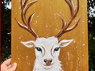 A hand holding a painting of a white reindeer with large antlers on a golden brown background. The image is decorated with colorful holiday lights and presents.