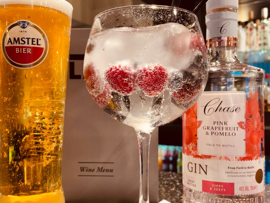 A glass of Amstel beer, a gin and tonic with raspberries and ice, and a bottle of Chase Pink Grapefruit & Pomelo Gin on a bar counter.