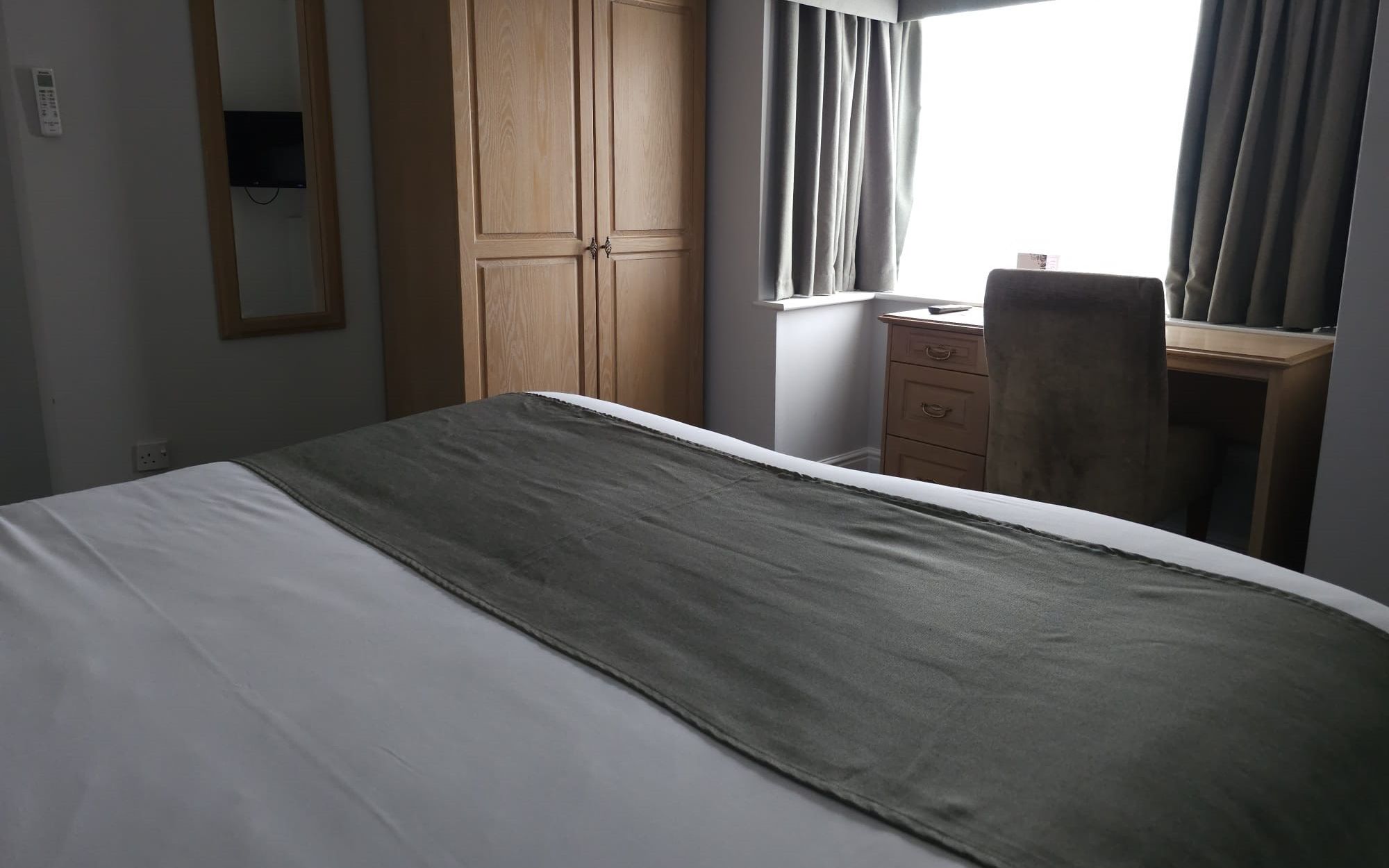 A tidy hotel room with a bed, wardrobe, mirror, desk, and chair near a window with curtains.