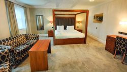Spacious hotel suite with a king-sized four-poster bed, modern wooden furniture, patterned sofa, and ample lighting.