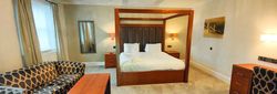 Spacious hotel suite with a king-sized four-poster bed, modern wooden furniture, patterned sofa, and ample lighting.