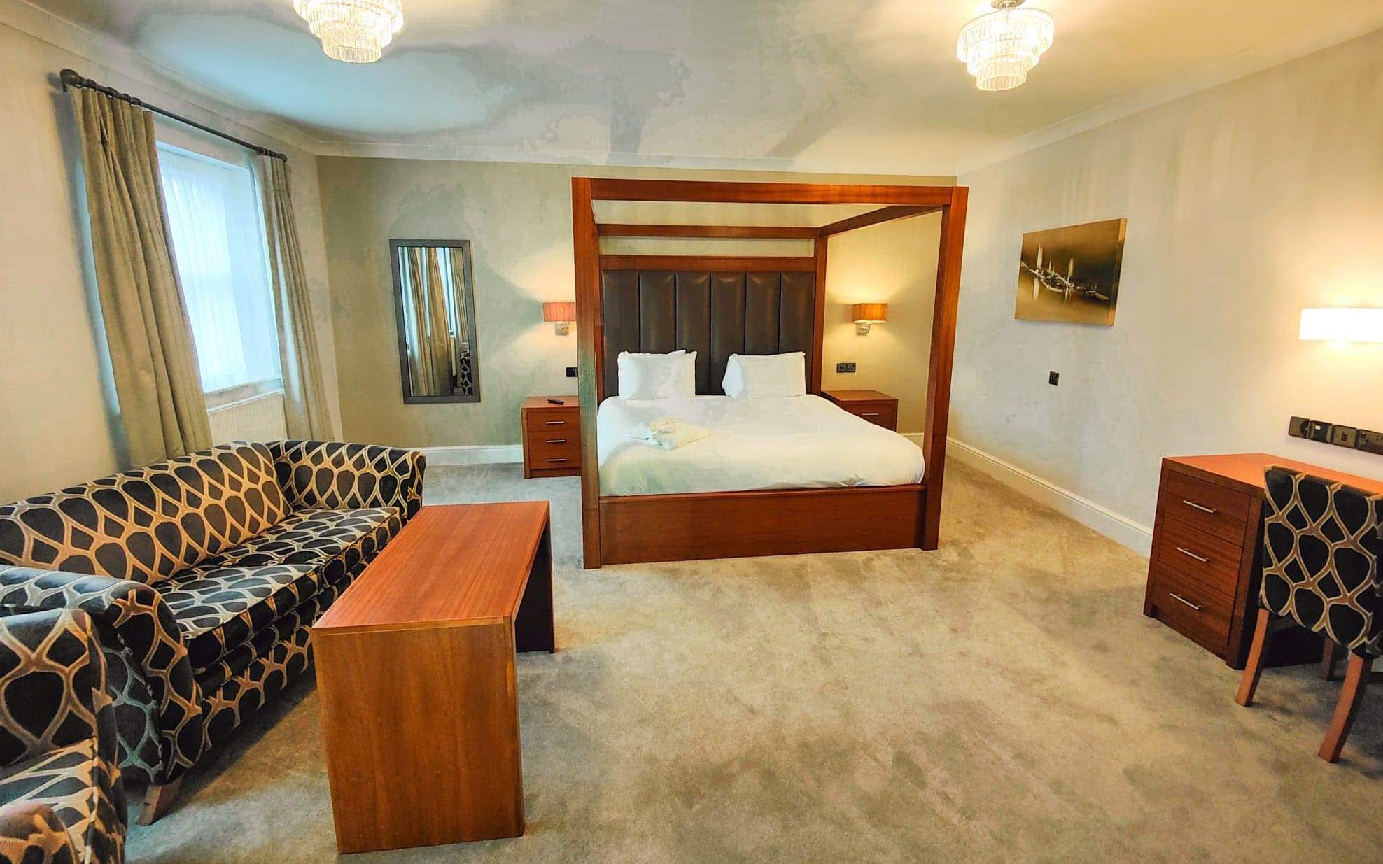 Spacious hotel suite with a king-sized four-poster bed, modern wooden furniture, patterned sofa, and ample lighting.