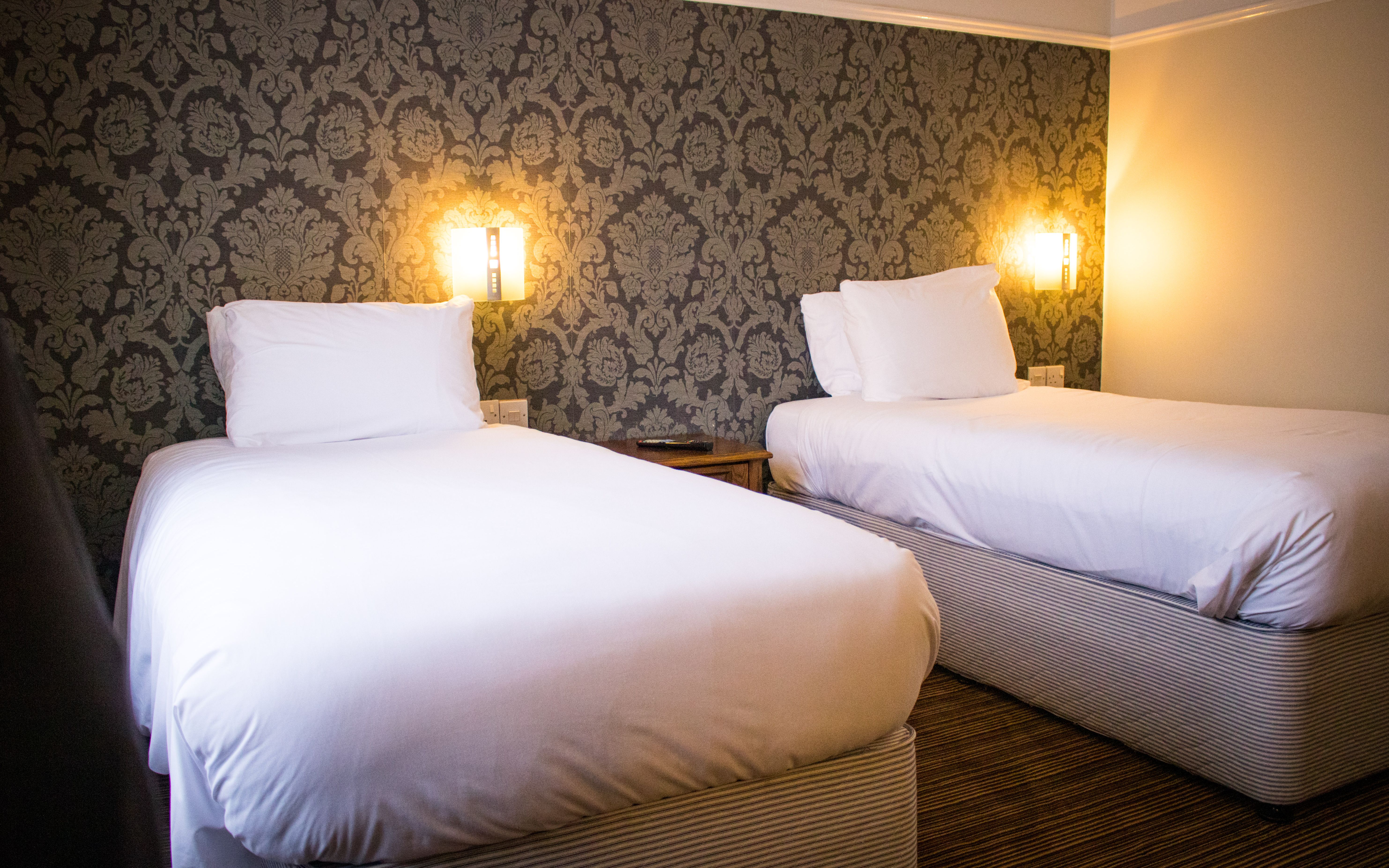 Two twin beds with white linens in a hotel room with patterned wallpaper and warm lighting.