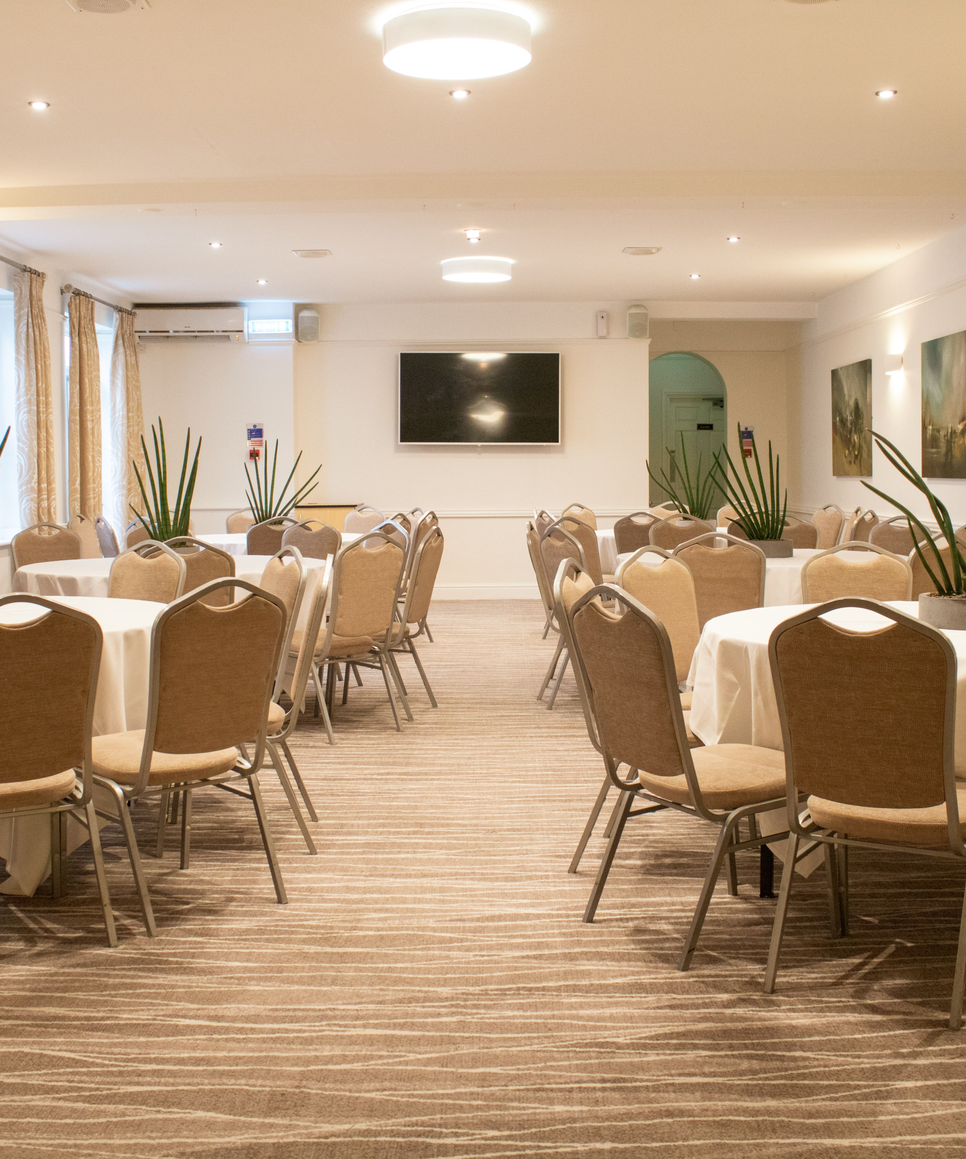 A conference room with round tables and beige chairs arranged neatly, decorated with potted plants.