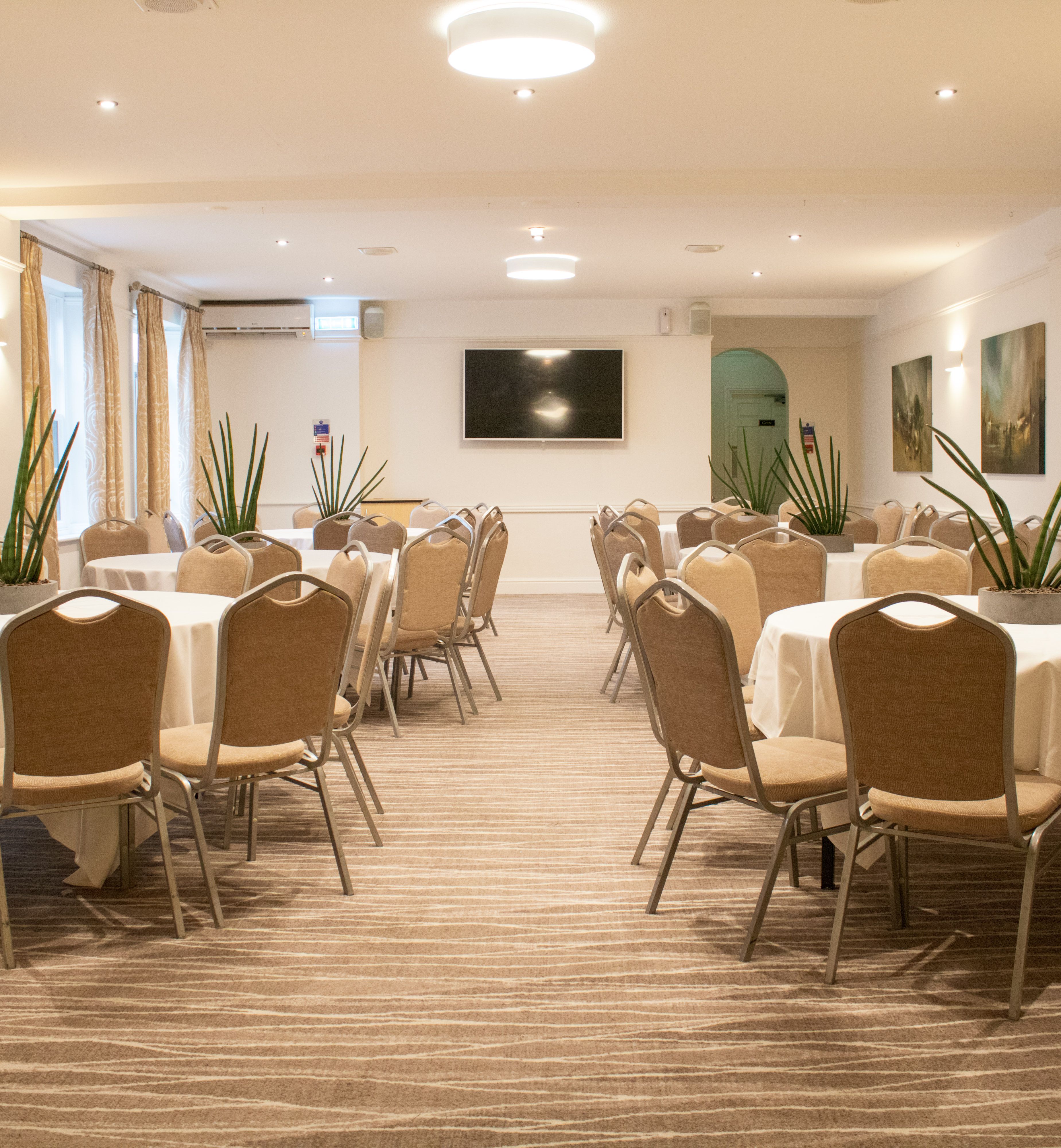 A conference room with round tables and beige chairs arranged neatly, decorated with potted plants.