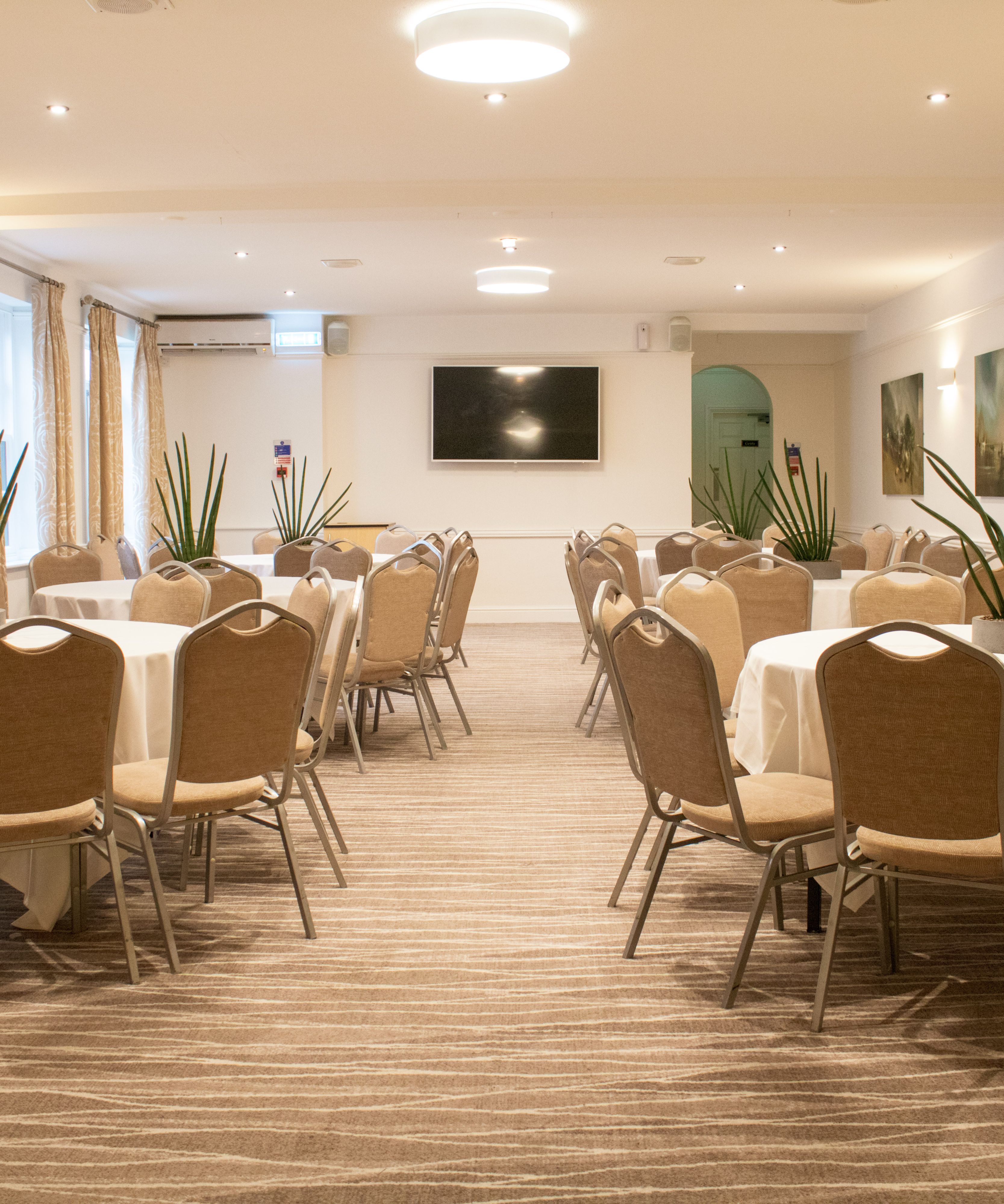 A conference room with round tables and beige chairs arranged neatly, decorated with potted plants.