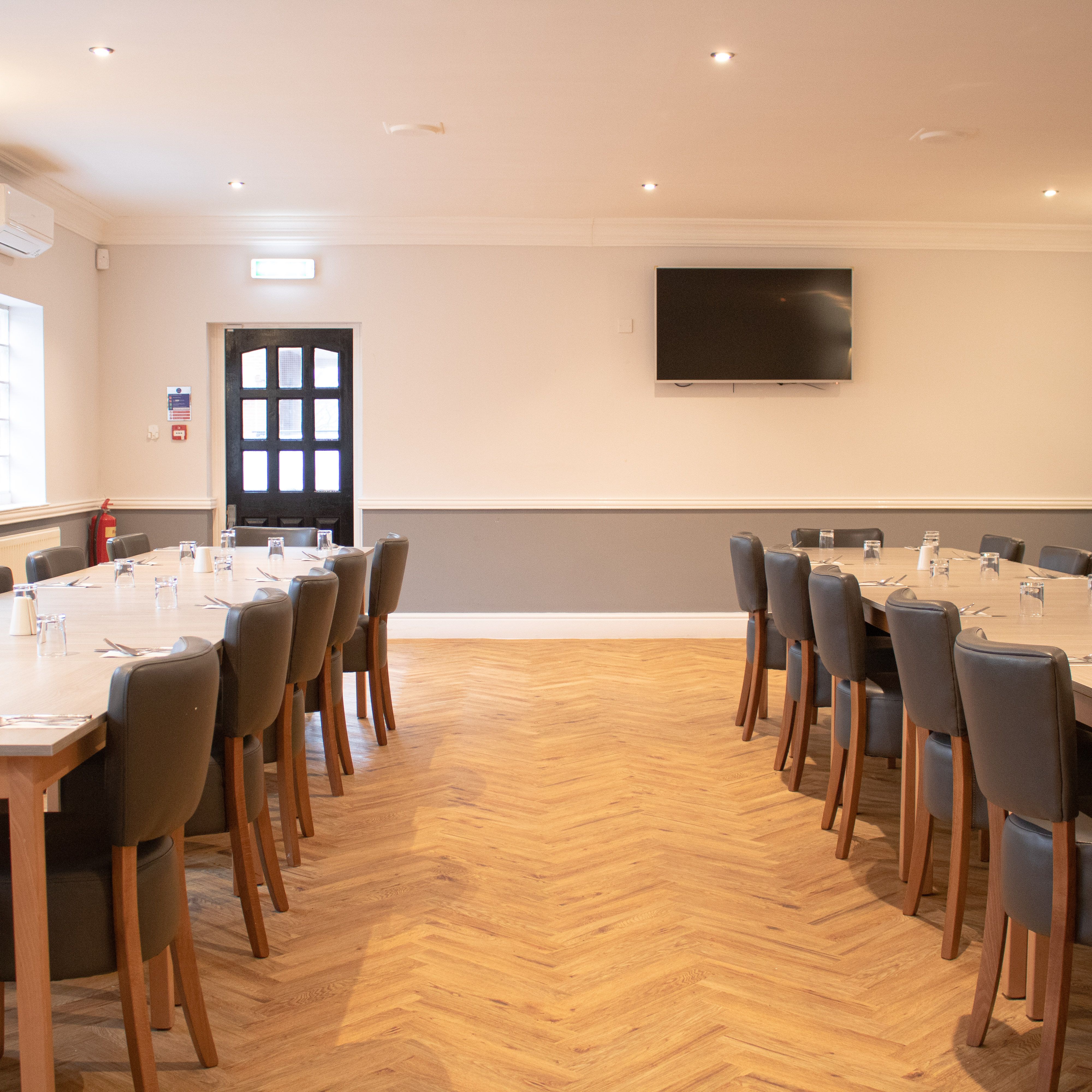 Spacious event room with two rows of tables, chairs, and place settings, featuring a television on the wall.