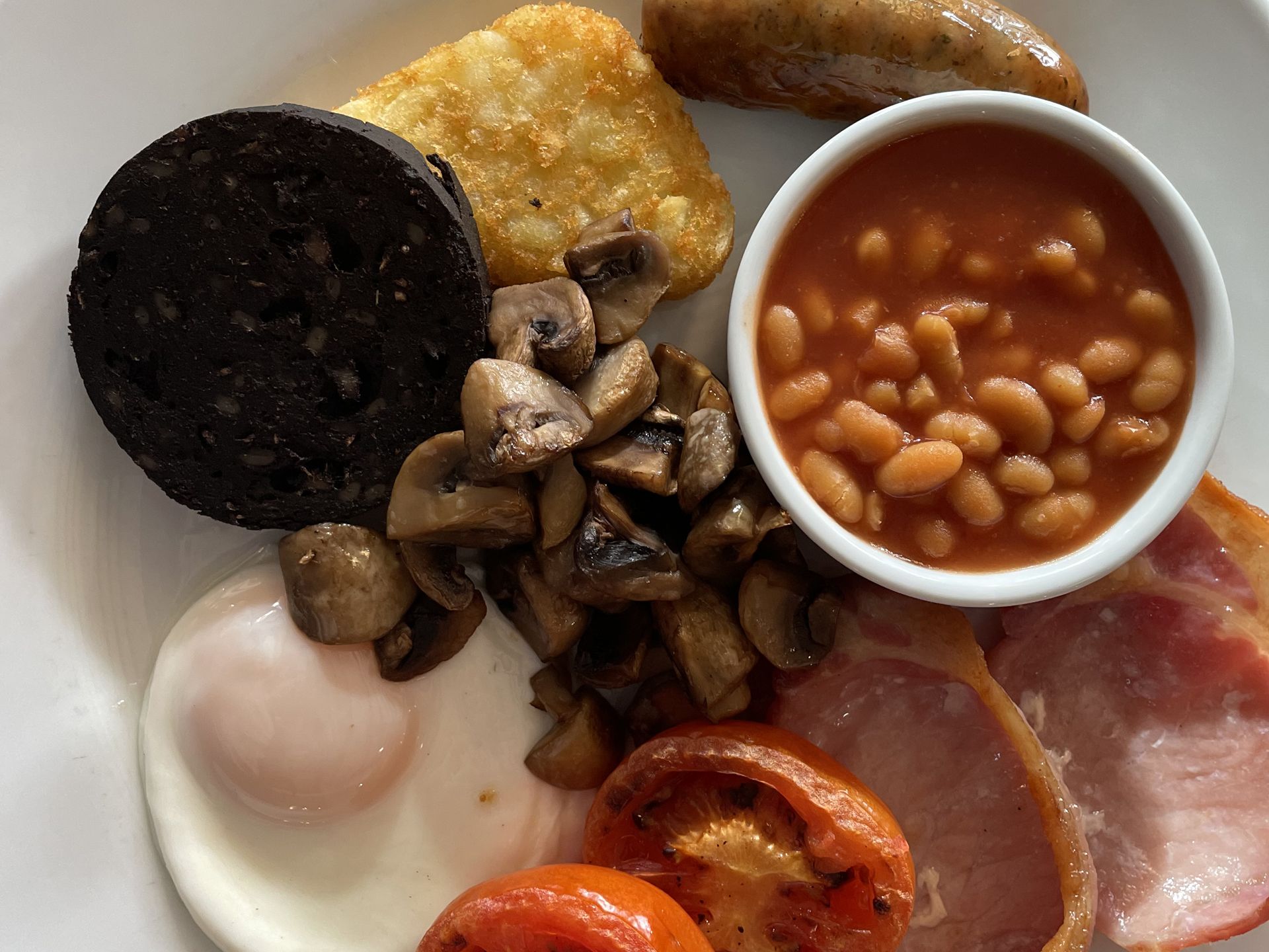 A plate of traditional English breakfast including fried egg, grilled tomato, bacon, sausage, hash brown, black pudding, sautéed mushrooms, and baked beans.