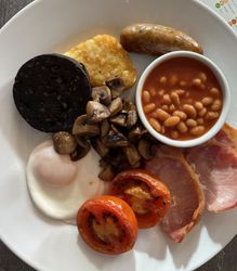A plate of traditional English breakfast including fried egg, grilled tomato, bacon, sausage, hash brown, black pudding, sautéed mushrooms, and baked beans.