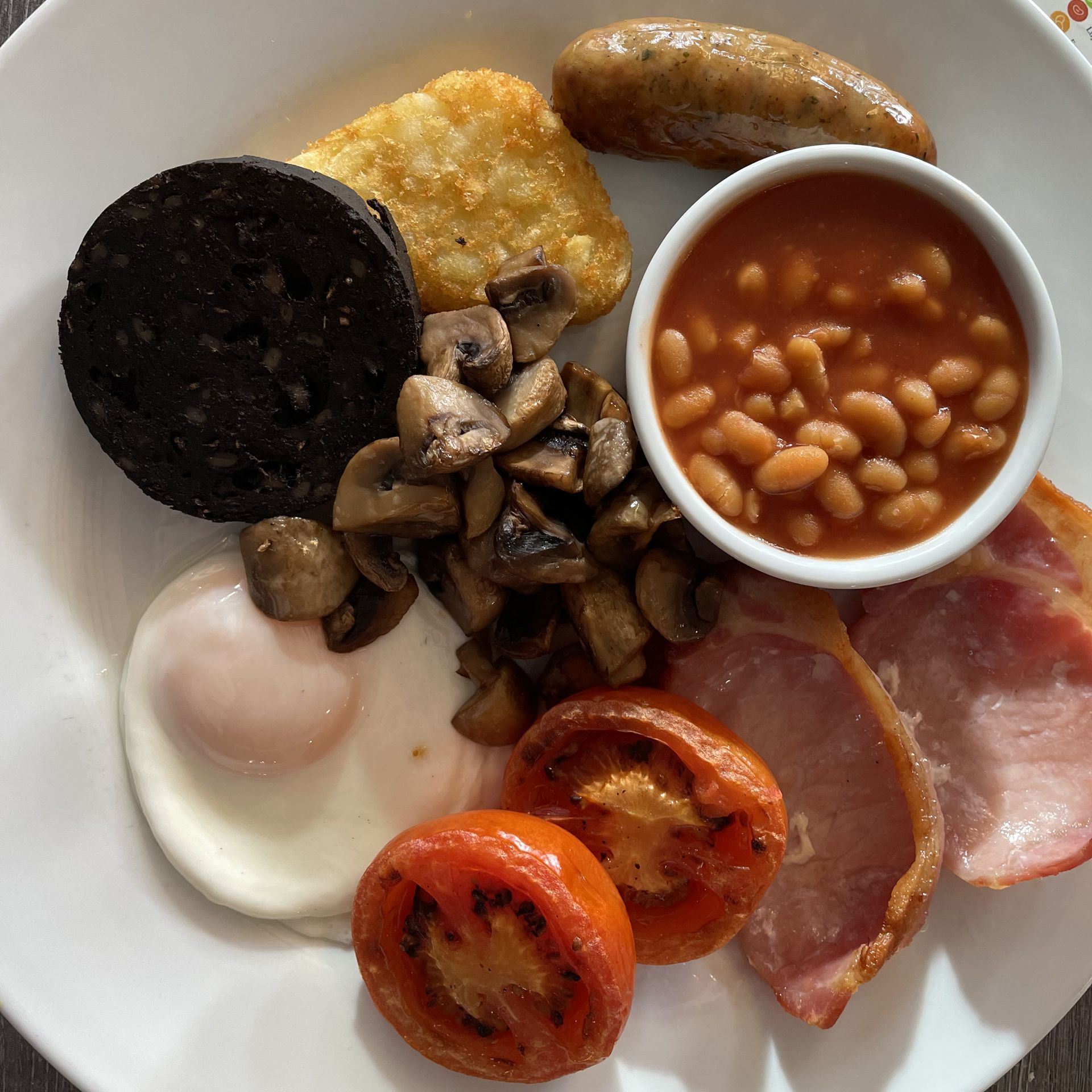 A plate of traditional English breakfast including fried egg, grilled tomato, bacon, sausage, hash brown, black pudding, sautéed mushrooms, and baked beans.