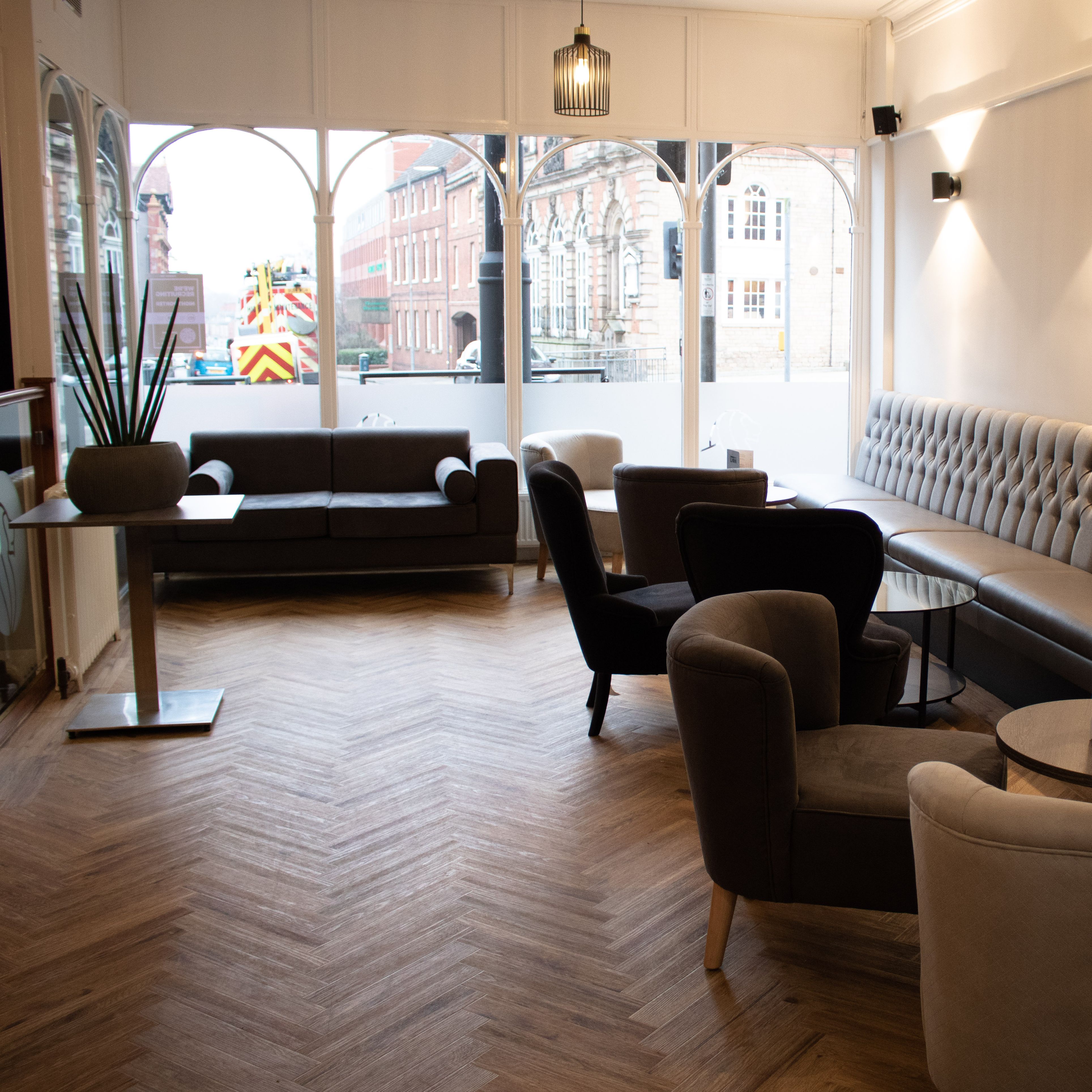 Modern lounge area in a restaurant with grey upholstered seating, round tables, and large windows.