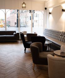 Modern lounge area in a restaurant with grey upholstered seating, round tables, and large windows.