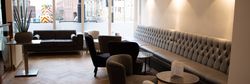 Modern lounge area in a restaurant with grey upholstered seating, round tables, and large windows.