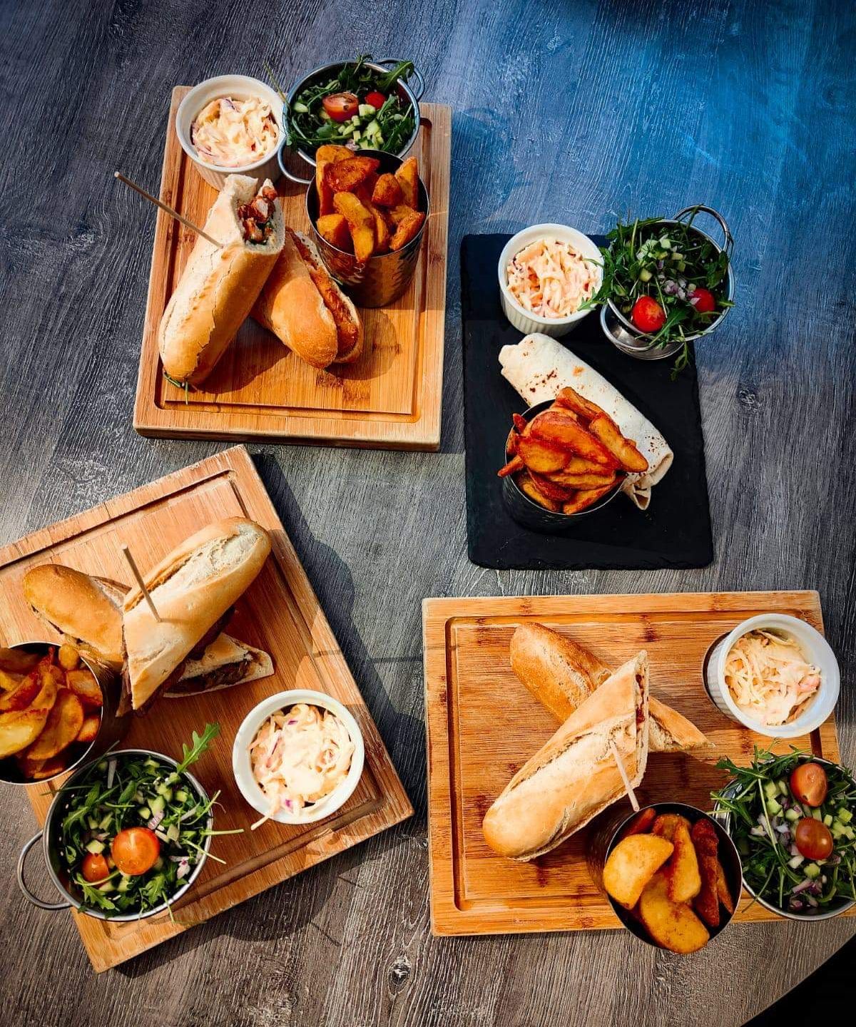 Assorted sandwiches served with potato wedges, coleslaw, and fresh salad on wooden boards.
