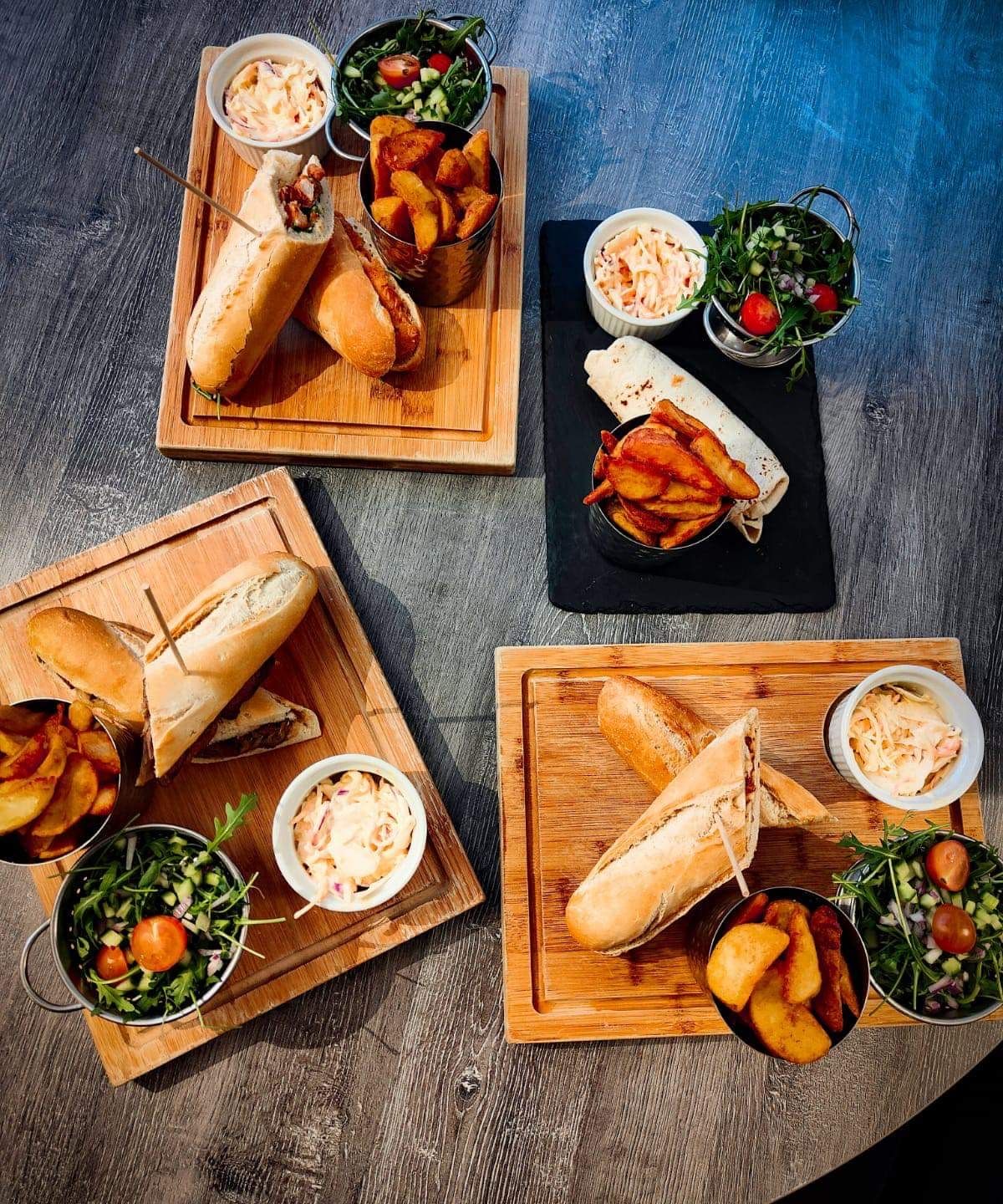 Assorted sandwiches served with potato wedges, coleslaw, and fresh salad on wooden boards.