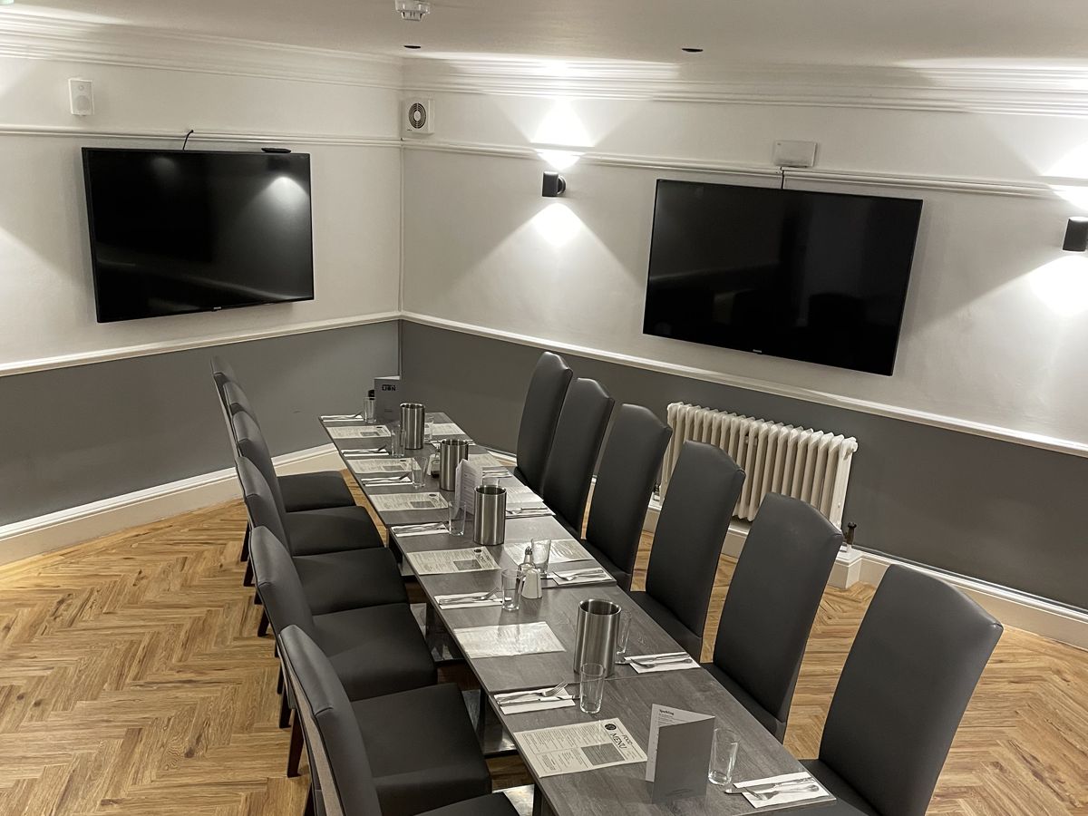 Modern dining room with a long table set for ten, grey chairs, and two flat-screen TVs on the wall.