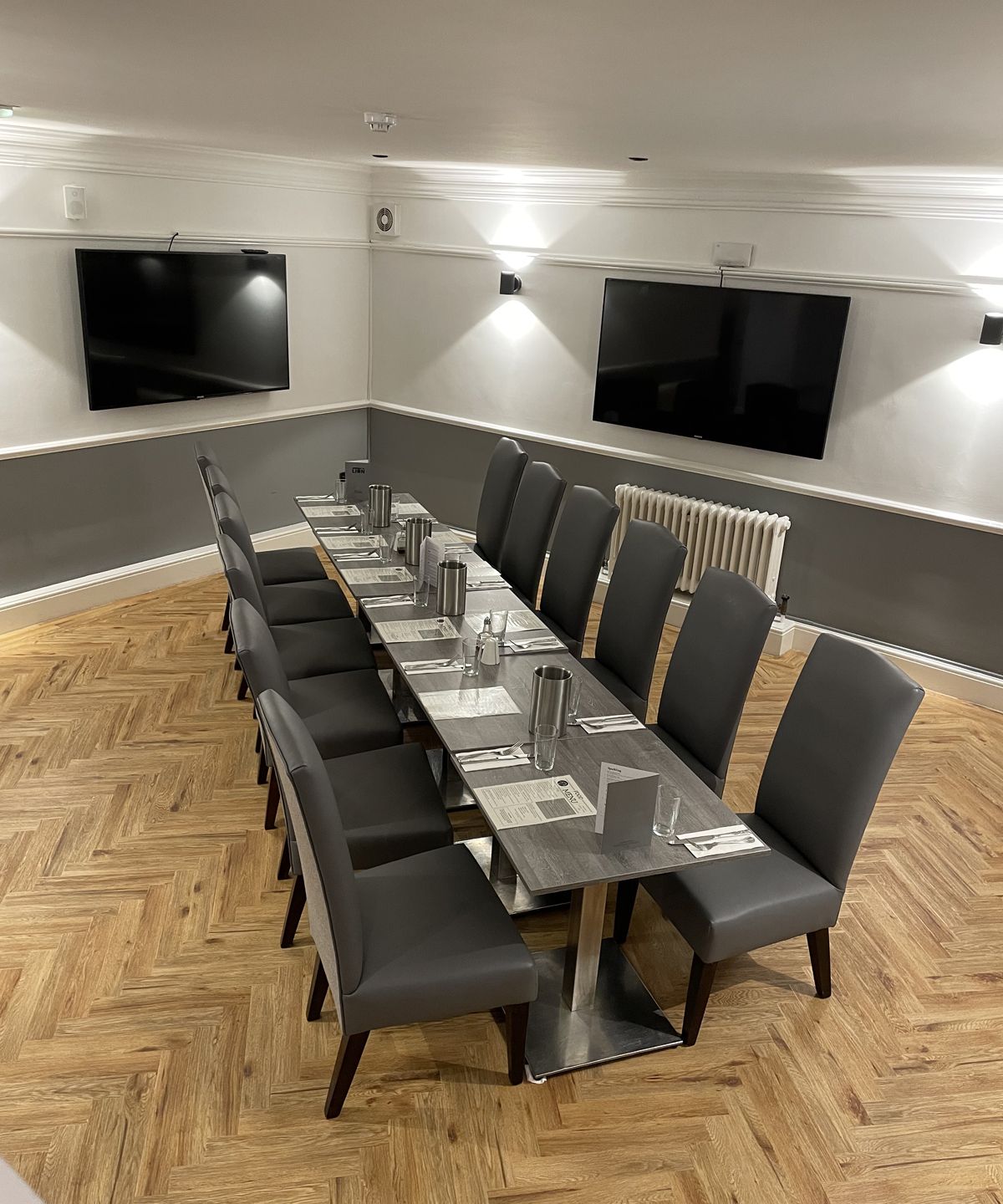 Modern dining room with a long table set for ten, grey chairs, and two flat-screen TVs on the wall.