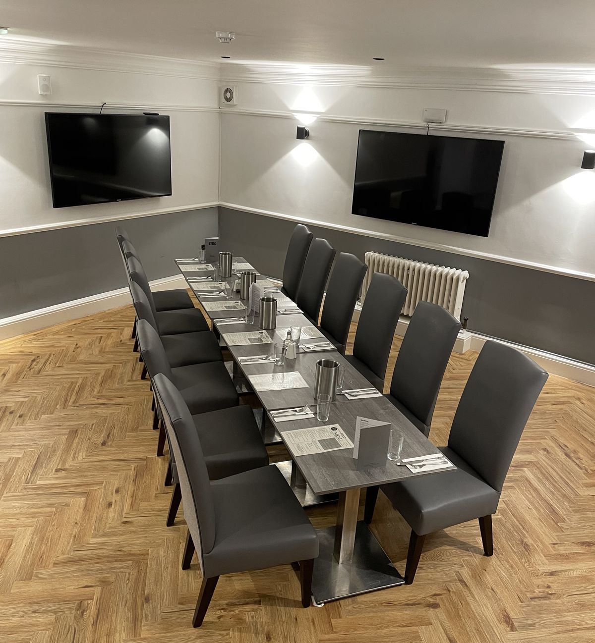 Modern dining room with a long table set for ten, grey chairs, and two flat-screen TVs on the wall.