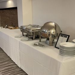 Buffet station set up in a conference room with serving trays, plates, and utensils on a long table covered with a white cloth.